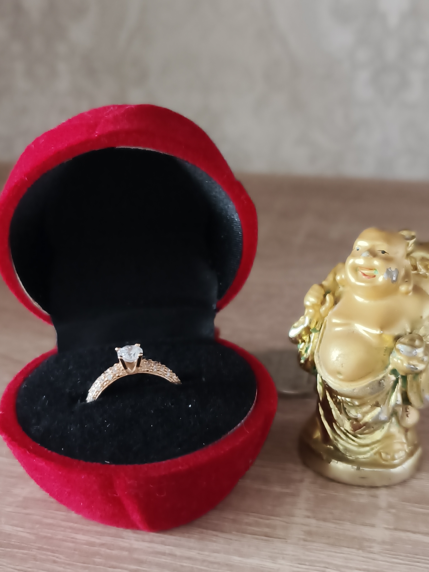 The image presents an indoor still life arrangement, likely captured in Kryvyi Rih, Ukraine. On a light brown wooden surface, a vibrant red, heart-shaped ring box is prominently featured on the left, open to reveal a black velvet interior. Inside the box, a gold-toned ring with a sparkling clear central gemstone, possibly a diamond or cubic zirconia, is elegantly displayed. The ring's band is also adorned with numerous smaller clear stones.
To the right of the ring box, a golden statuette of a laughing Buddha, or Hotei, stands. The Buddha figure is depicted with a jovial expression, a large belly, and a wide smile, symbolizing happiness and prosperity. The statuette appears to be made of a painted material, showing some slight wear and imperfections in its gold finish, with hints of green and blue coloring visible in areas.
The wooden surface exhibits a subtle grain pattern, and the background is a soft-focus wall, light in color, with a faint, repeating textured pattern. The lighting suggests an ambient indoor setting. No specific activity is depicted, but the presence of the engagement-style ring and the symbol of good fortune from the Buddha statuette collectively evoke themes of happiness, commitment, and positive life events. There is no visible text in the image.