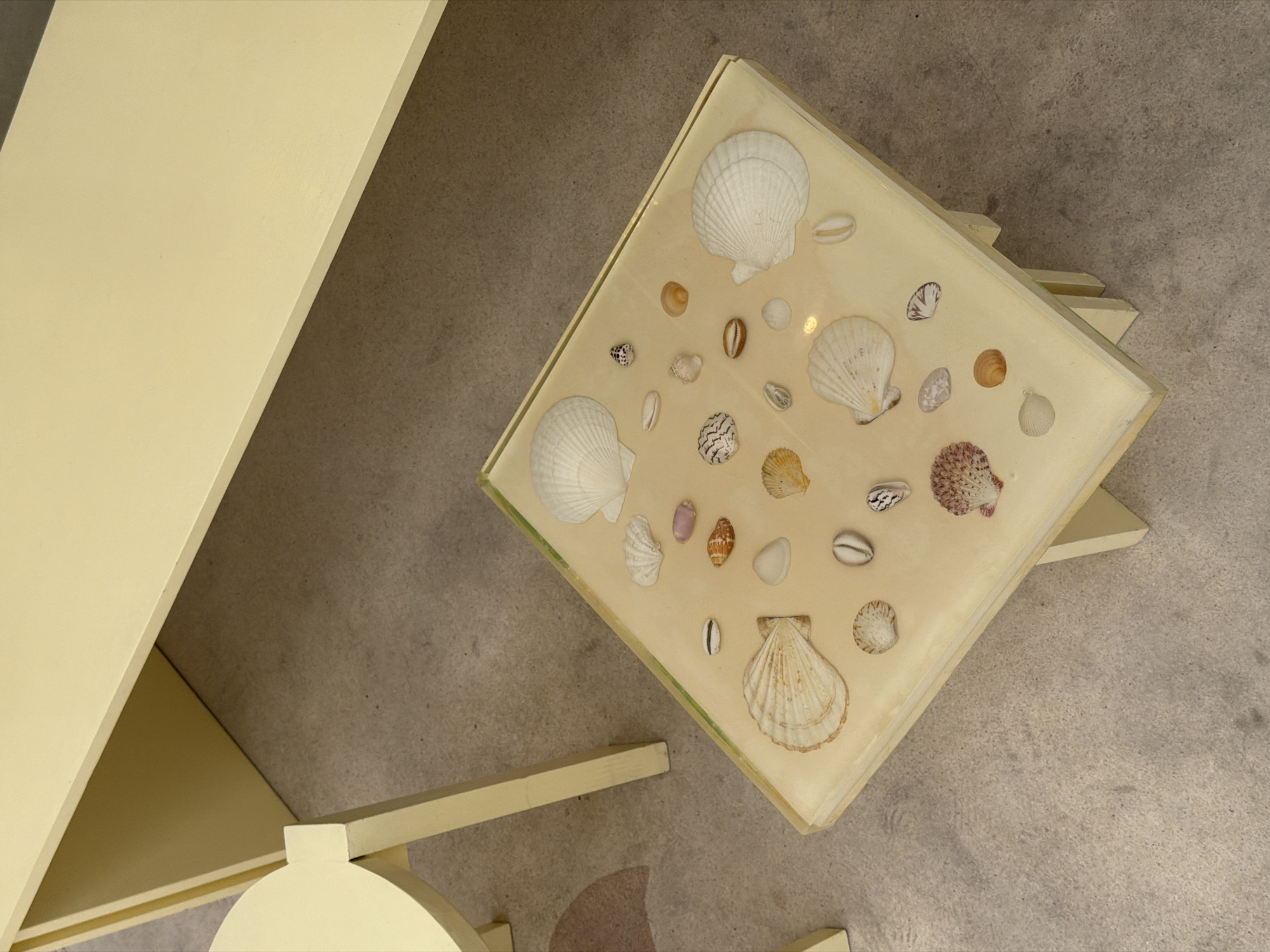 In Mengwi, Indonesia, a light yellow square side table holds a collection of seashells embedded in a clear resin top. The table has four light yellow legs. To the left of the table, a portion of a round, light yellow stool is visible, with two legs. A larger, light yellow table or surface extends from the upper right of the frame. The floor beneath and around the furniture appears to be concrete. The overall scene is brightly lit, suggesting an indoor or shaded outdoor setting.