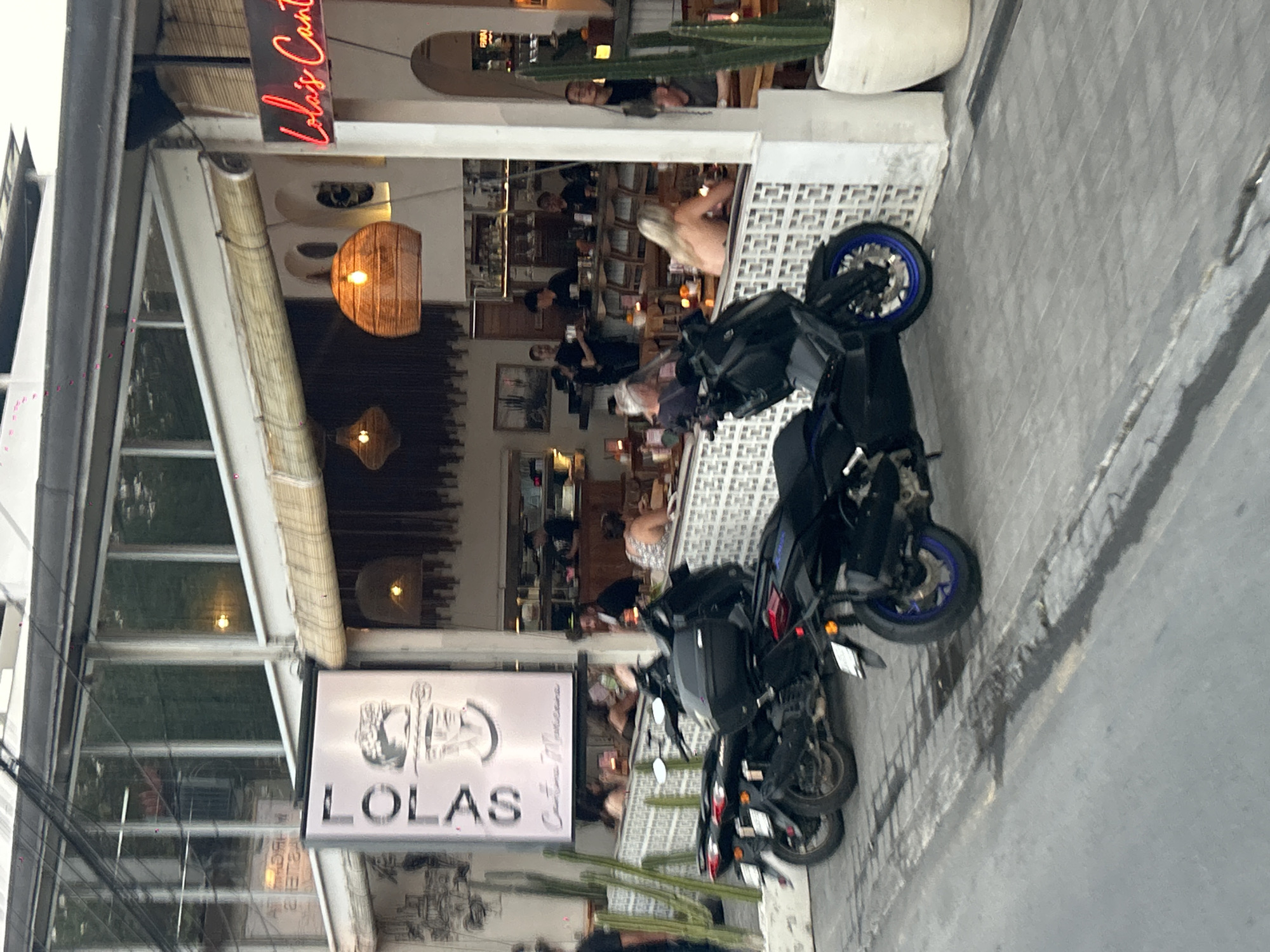 In North Kuta, Indonesia, a street scene unfolds in front of a restaurant named "Lola's Cantina". Several parked motorcycles, primarily scooters, line the paved street. The restaurant features a rustic aesthetic with bamboo blinds, woven light fixtures, and white patterned wall accents. Inside, patrons are seated at tables, some interacting with each other and others appearing to converse with staff behind a bar area. One sign prominently displays "LOLA'S" in neon pink, and another, a vertical sign, reads "LOLA'S Cantina Mexicana". Cacti in pots are visible near the entrance, adding to the establishment's ambiance. The overall atmosphere suggests a lively dining or social setting.