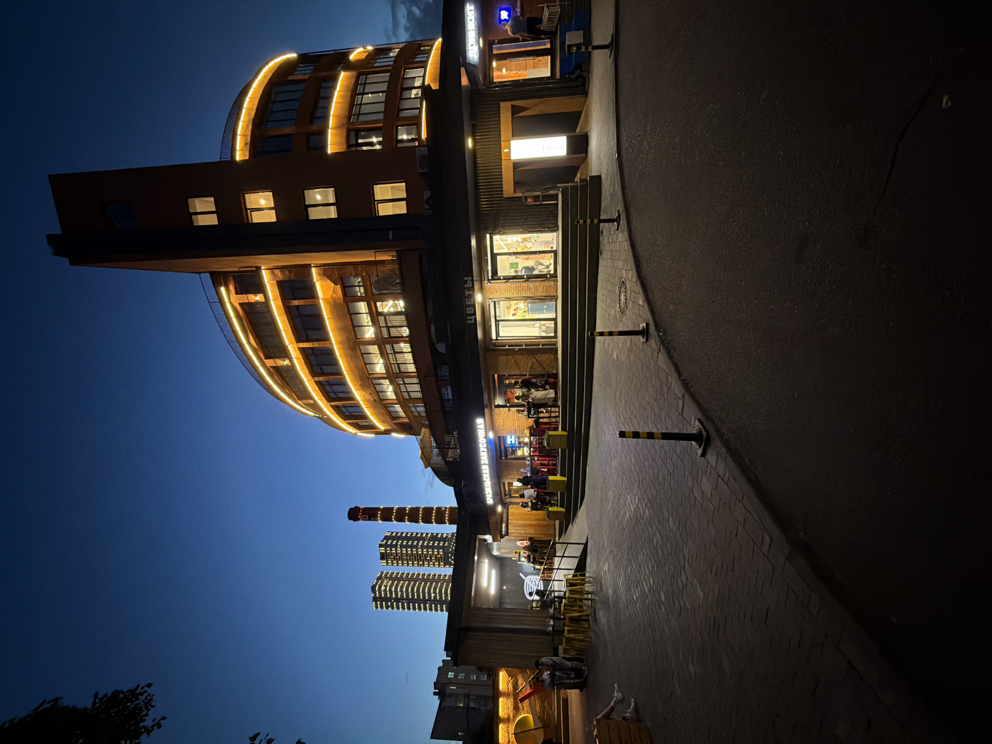 The image, taken at dusk or night in Khimki, Russia, presents a vibrant urban scene dominated by a large, modern building with a curved, multi-story facade. The building is extensively illuminated with warm yellow lights outlining its architecture and emanating from its numerous windows, casting a warm glow.
At the base of this main building, several commercial establishments are active, featuring outdoor seating areas where people are visible, likely dining or socializing. The storefronts are brightly lit, contrasting with the deep blue twilight sky.
In the background, two tall, slender, industrial-style structures, possibly chimneys or repurposed towers, stand against the dark sky. One of these structures is adorned with vertical lines of lights, and other distant buildings are also visible, hinting at a developed urban landscape.
The foreground shows a paved walkway or street with bollards marked with yellow and black stripes. Small trees or foliage can be seen on the far left.
Visible text includes "ЦВЕТЫ" (Flowers) above one shop and "ВЫПЕЧКА ЗАКУСОЧНАЯ" (Bakery Snack Bar / Baked Goods Eatery) on another sign. Another partially visible sign appears to read "МОЛОКО" (Milk). The overall atmosphere is lively and inviting, characteristic of an urban area in the evening.