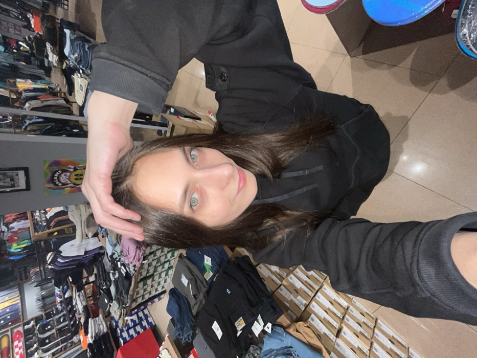 A young adult female, with long dark hair and light green eyes, captures a high-angle selfie inside the BALANCE streetwear and skate shop located at Mira Avenue 94, Krasnoyarsk, Russia. She wears a black hooded sweatshirt with a distinctive circular patch and buttons on the left sleeve. Her left hand is positioned on the top of her head, and she displays a slight smile. The retail environment features numerous displays of folded clothing, including items from Polar Skate Co., stacked shoeboxes, and vertically mounted skateboard decks against a back wall. A prominent colorful poster depicting a yellow cartoon smiley face and the text "BALANCE BBQ" is visible on the rear wall. The floor is composed of light-colored tiles.