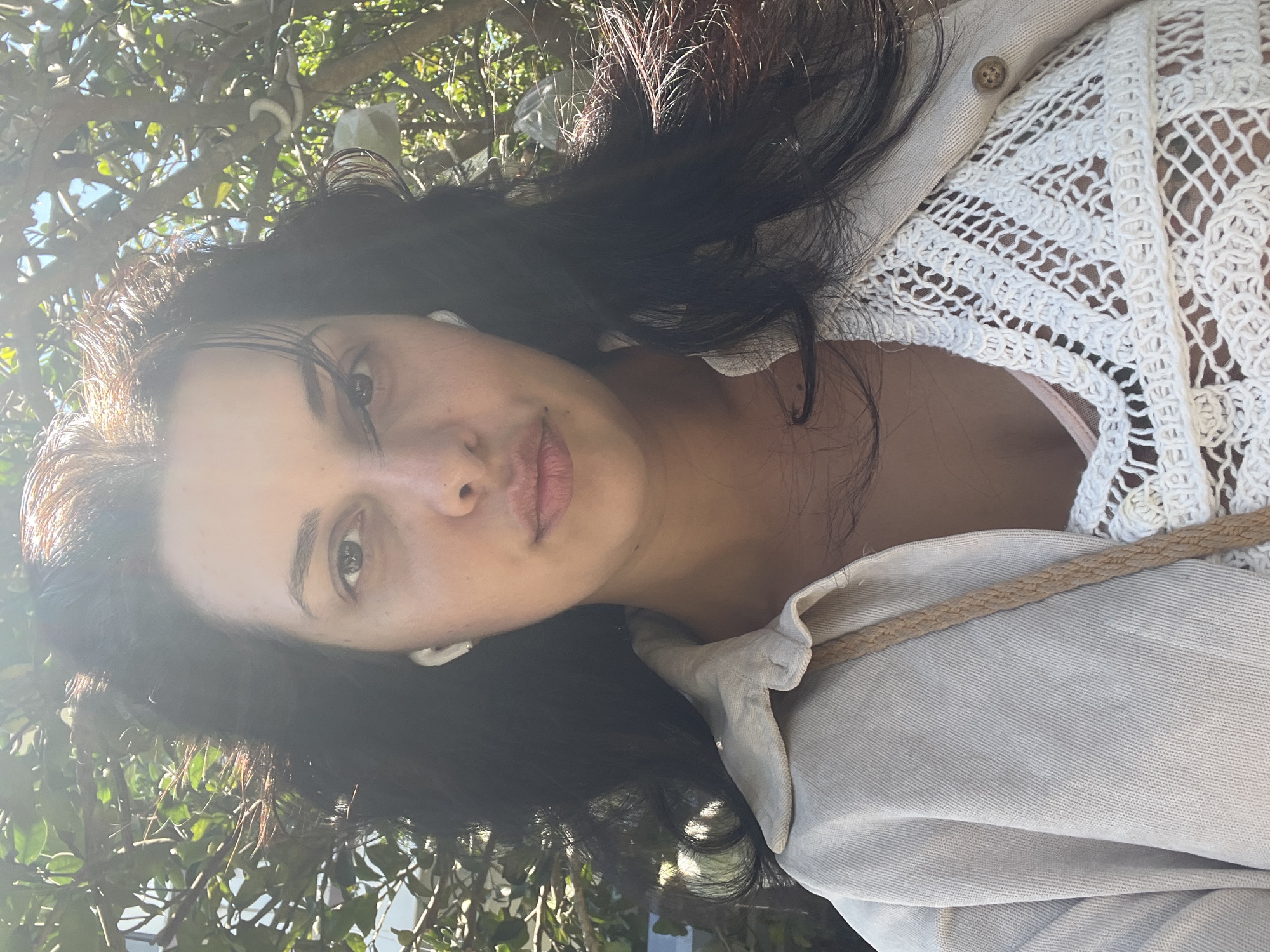 A young woman with long, dark, wavy hair and light brown eyes is shown in a close-up, selfie-style shot. She is outdoors, with a backdrop of green foliage and branches, suggesting a park or natural area. The lighting indicates it is daytime, with sunlight casting shadows and highlights on her face and hair. She is wearing a light-colored collared jacket over a white, crocheted top. White wireless earbuds are visible in her ears. Her expression is neutral.
There is no information in the image to indicate a specific location within Đà Nẵng, Vietnam. The scene is generic and could be found in many locations with similar vegetation and lighting.