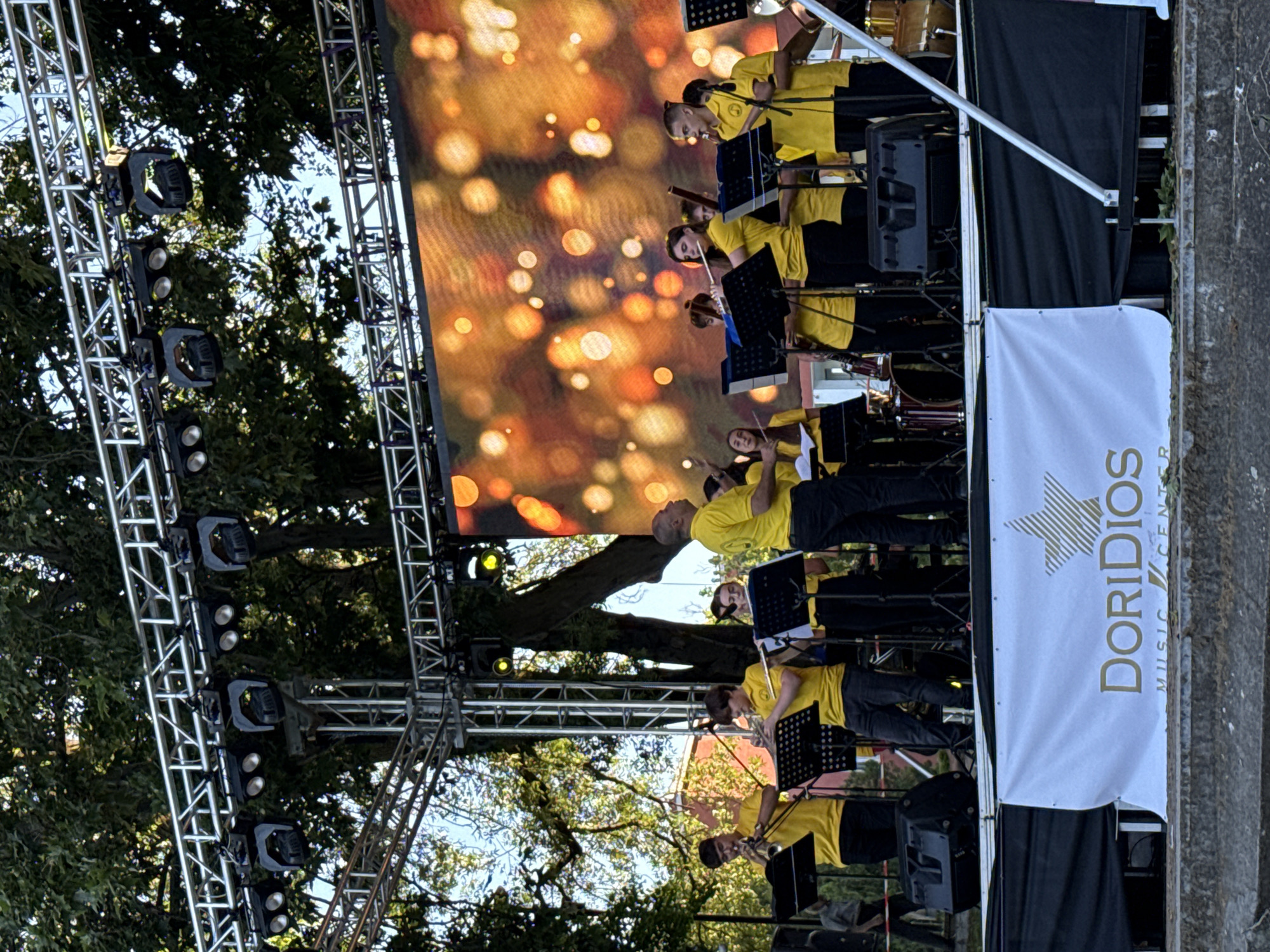 An outdoor stage with a band performing is set up in Pleven, Bulgaria, possibly in a park. The band members, all wearing bright yellow shirts, are playing various instruments including trumpets and flutes. They are positioned on a raised stage in front of a large LED screen displaying a blurry, bokeh-like pattern of orange and yellow lights. Stage lighting rigs are mounted above the performers, and large speakers are placed on the stage. A white banner with the text "DORIDIOS MUSIC CENTER" is visible at the front of the stage. The backdrop behind the stage features trees, suggesting a natural, outdoor setting within the city.
