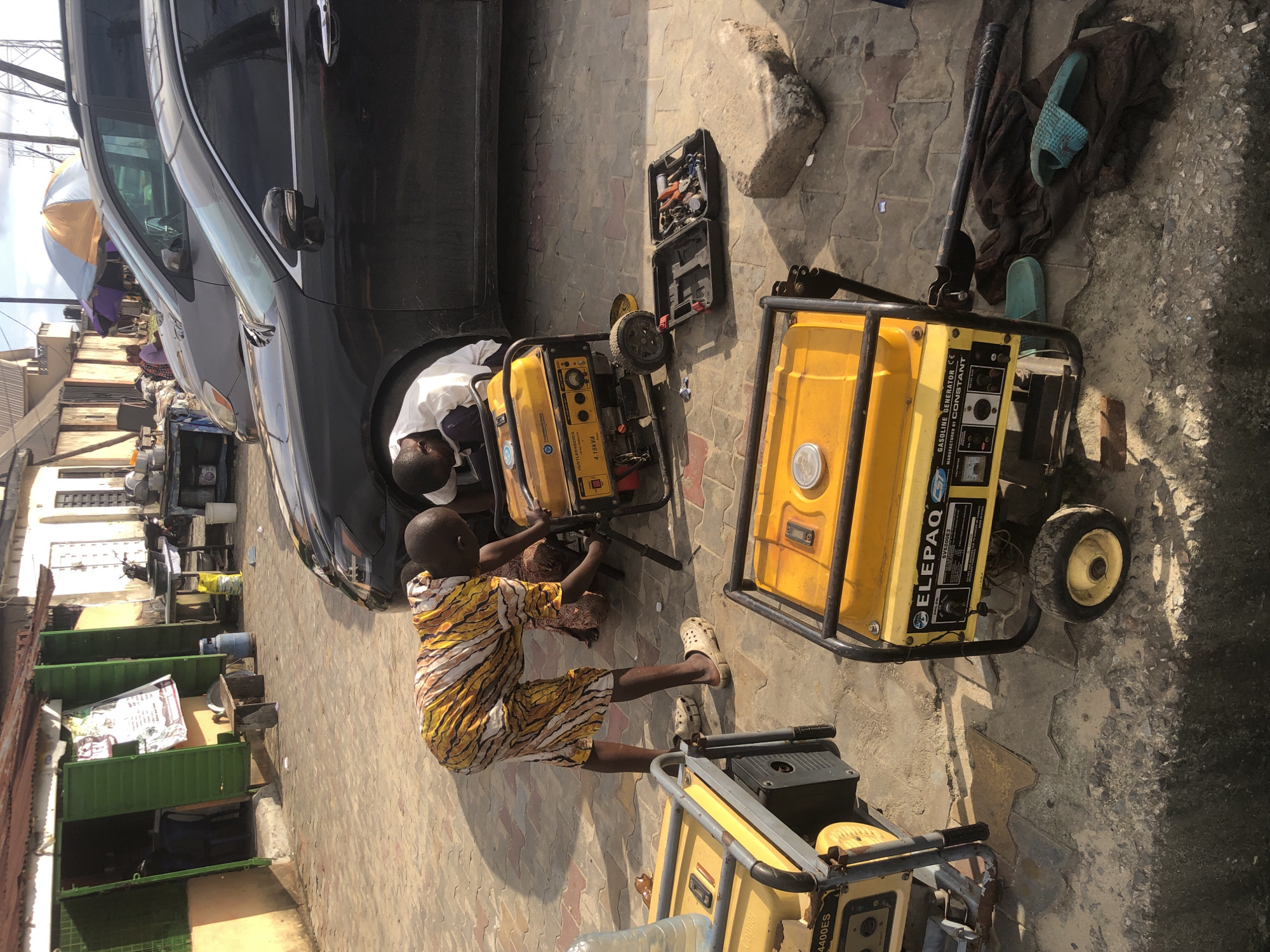 In Ado, Nigeria, two individuals are observed tending to a yellow portable generator situated on a tiled pavement. One person, wearing a colorful patterned shirt and shorts, bends over the generator. The other individual, dressed in a white shirt, is partially obscured by the generator. Nearby, a black tool case lies open, revealing various tools. Two other yellow generators are visible, one to the left and another in the foreground. A black sedan is parked behind the people and generators. The background shows a row of buildings with storefronts and a grey van. The scene appears to be an outdoor setting, possibly a street or market area.