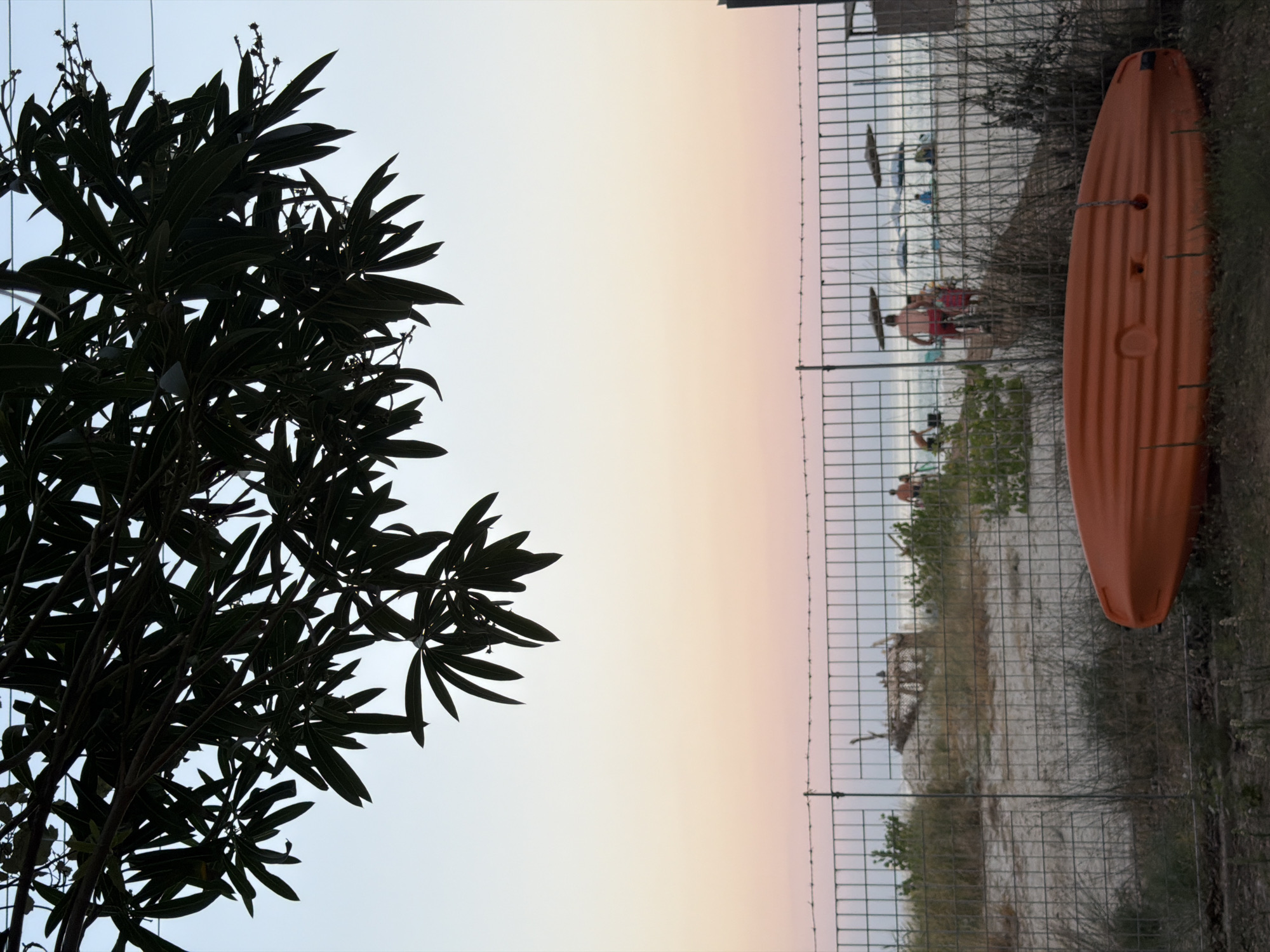 An outdoor scene depicts a beach with a fence in the foreground and trees overhead. The sky transitions from a light blue at the top to soft hues of pink and orange near the horizon, indicating either sunrise or sunset. In the foreground, an orange kayak lies on the ground. A wire mesh fence runs across the middle ground, separating the viewer from the beach. Behind the fence, people are visible on the sandy beach, some relaxing under umbrellas. The ocean stretches out in the background. The overall impression is one of a peaceful, coastal setting.