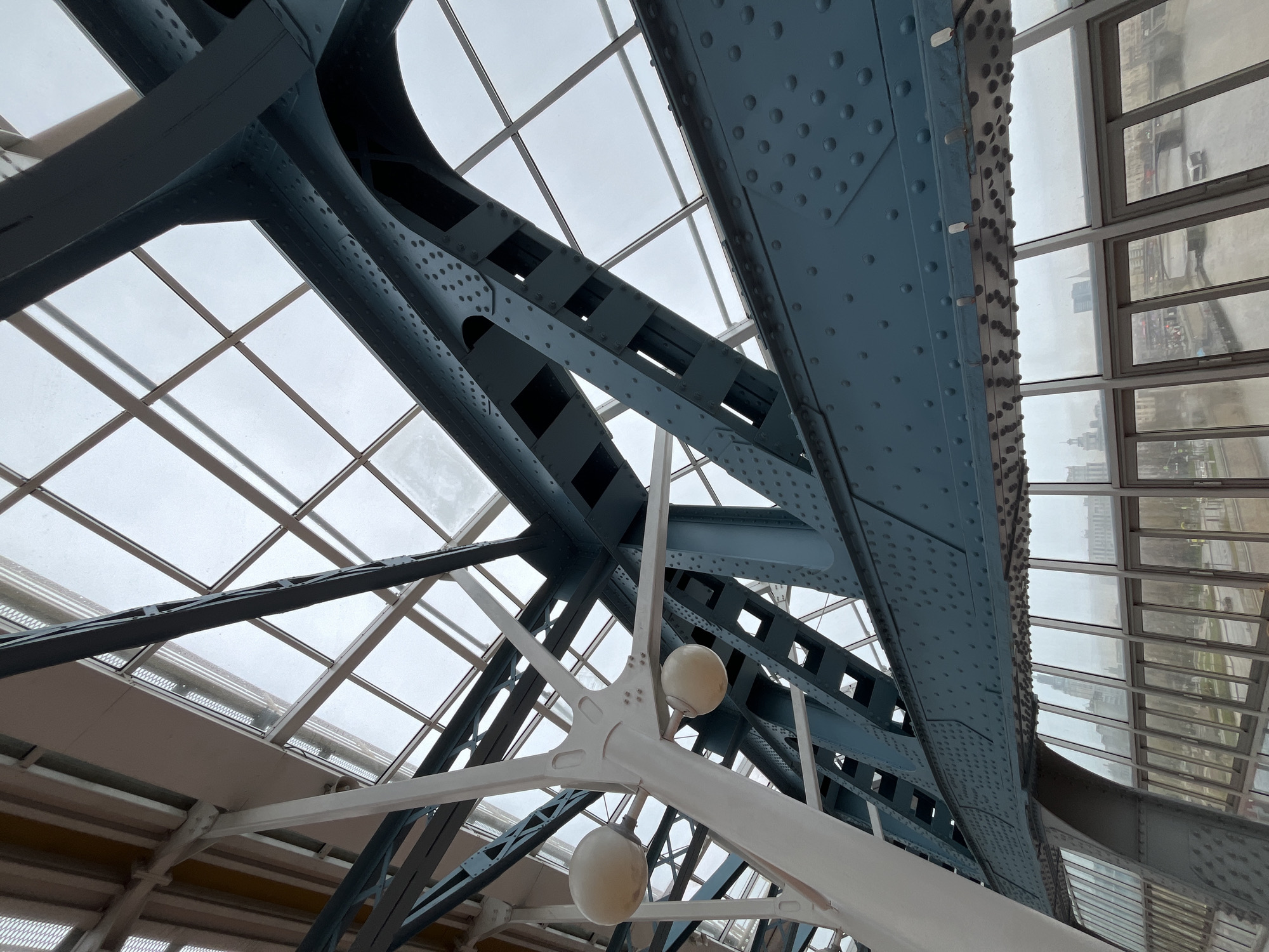 The image shows an upward view of the interior structure of the Krymsky Bridge in Moscow, Russia. Visible are the bridge's blue steel trusses secured with rivets. A glass ceiling overhead provides diffused daylight. White structural supports with spherical lamp fixtures are positioned beneath the trusses. A window wall on the right side provides a view of the city. The sky is overcast.