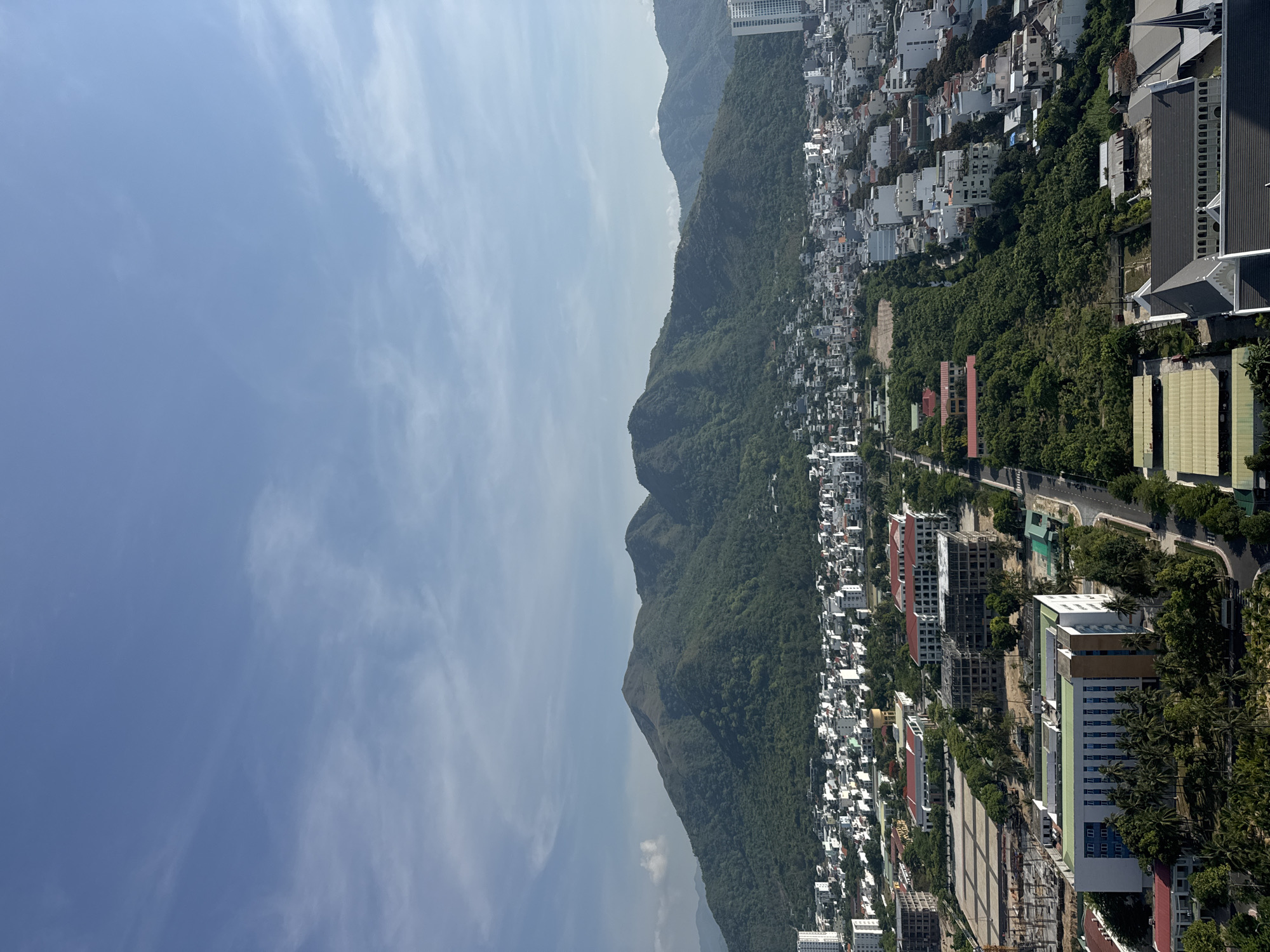 This is a high-angle aerial photograph taken on a clear day with a few wispy clouds in the sky. The image captures a sprawling city with a backdrop of lush green mountains. In the foreground, there are several buildings, including some under construction, and a road winding through the urban landscape. The midground shows a dense residential area with white and beige buildings, and further back, the imposing, tree-covered mountains dominate the horizon. The lighting suggests it is daytime, likely mid-morning or afternoon. There are no people visible in the image. The location is described as Unknown, Unknown.