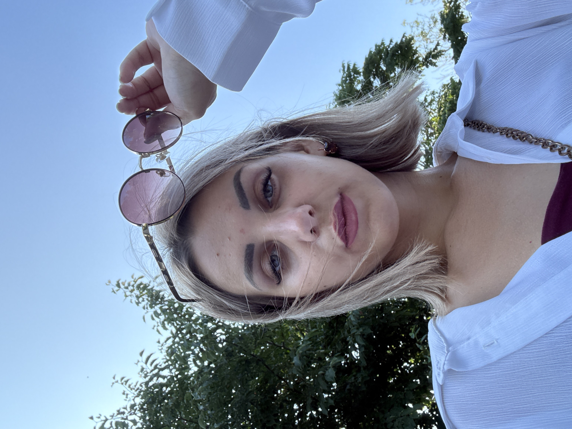 A close-up, low-angle selfie shows a young Caucasian woman with blonde bobbed hair and light makeup, holding a pair of round, pink-tinted sunglasses above her head with her right hand. She is wearing a white, sheer button-up shirt over a maroon garment and a gold chain strap of a bag is visible across her chest. Her face is slightly tilted, and she is looking directly at the camera. The background consists of a clear blue sky and the foliage of a green tree. The scene appears to be outdoors during the daytime. This image was taken in Yuzhno-Sakhalinsk, Russia.