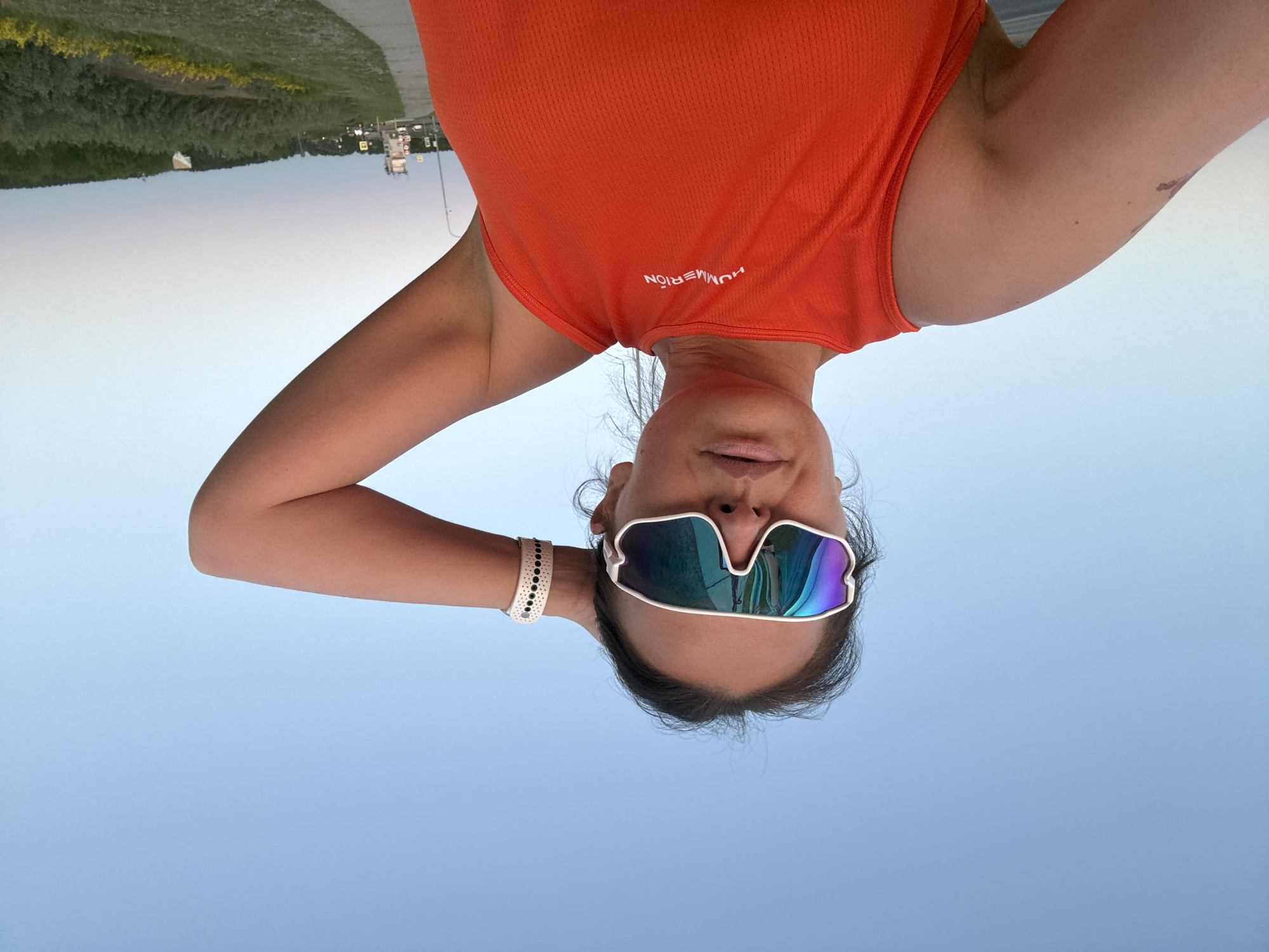 A woman is pictured in a low-angle selfie with an upside-down orientation. She is wearing an orange tank top and sunglasses with blue and purple lenses. She has her arms behind her head, and is wearing a bracelet. A landscape with trees and a road is in the background. The photo appears to be taken outdoors. There is no indication that the photo was taken in the city of Chaikovsky, Russia.