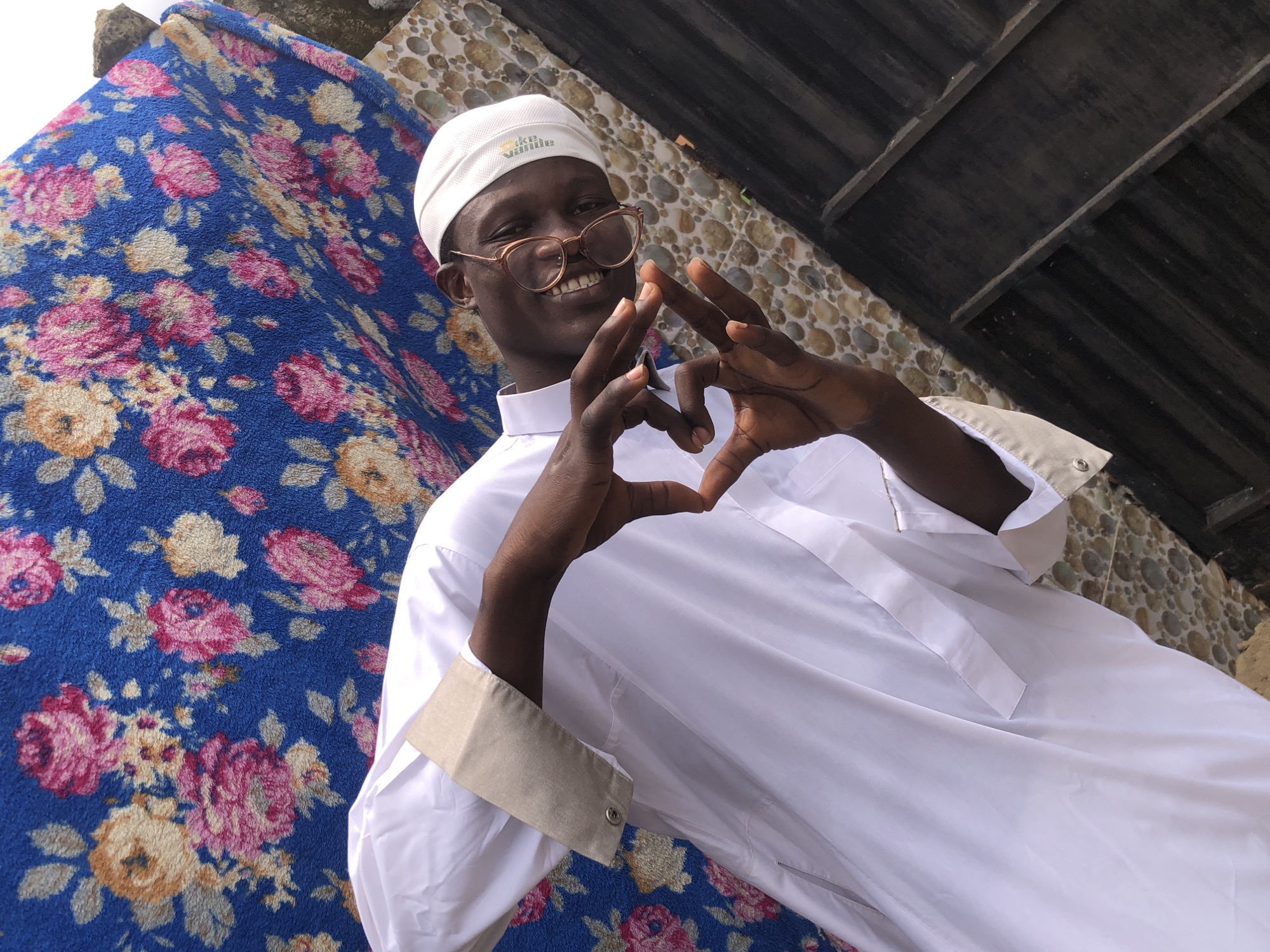 In Ado, Nigeria, a young man in a white thawb and a white kufi hat poses for a photo. He is wearing round glasses and smiling, his head tilted. His hands are held up, forming a heart shape. The background features a blue floral tapestry draped over a textured stone wall and a dark wooden structure.