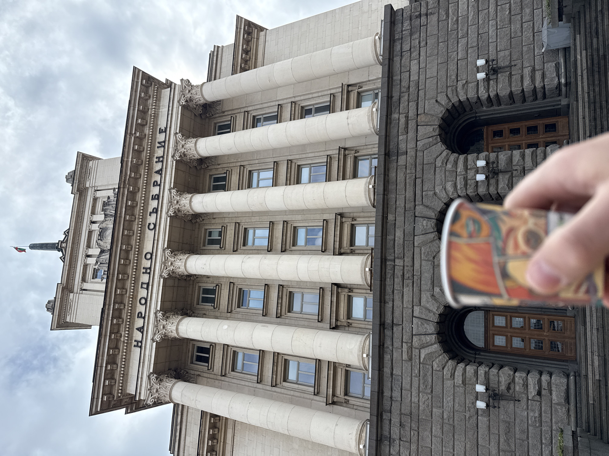The image depicts the National Assembly of Bulgaria in Sofia. The building is a large, imposing structure with a facade of light-colored stone columns and a darker, gray stone facade to the right. The letters "НАРОДНО СЪБРАНИЕ" are displayed vertically along the facade. Windows are interspersed between the columns. The sky is overcast. A blurred hand holding an object is partially visible in the foreground.
