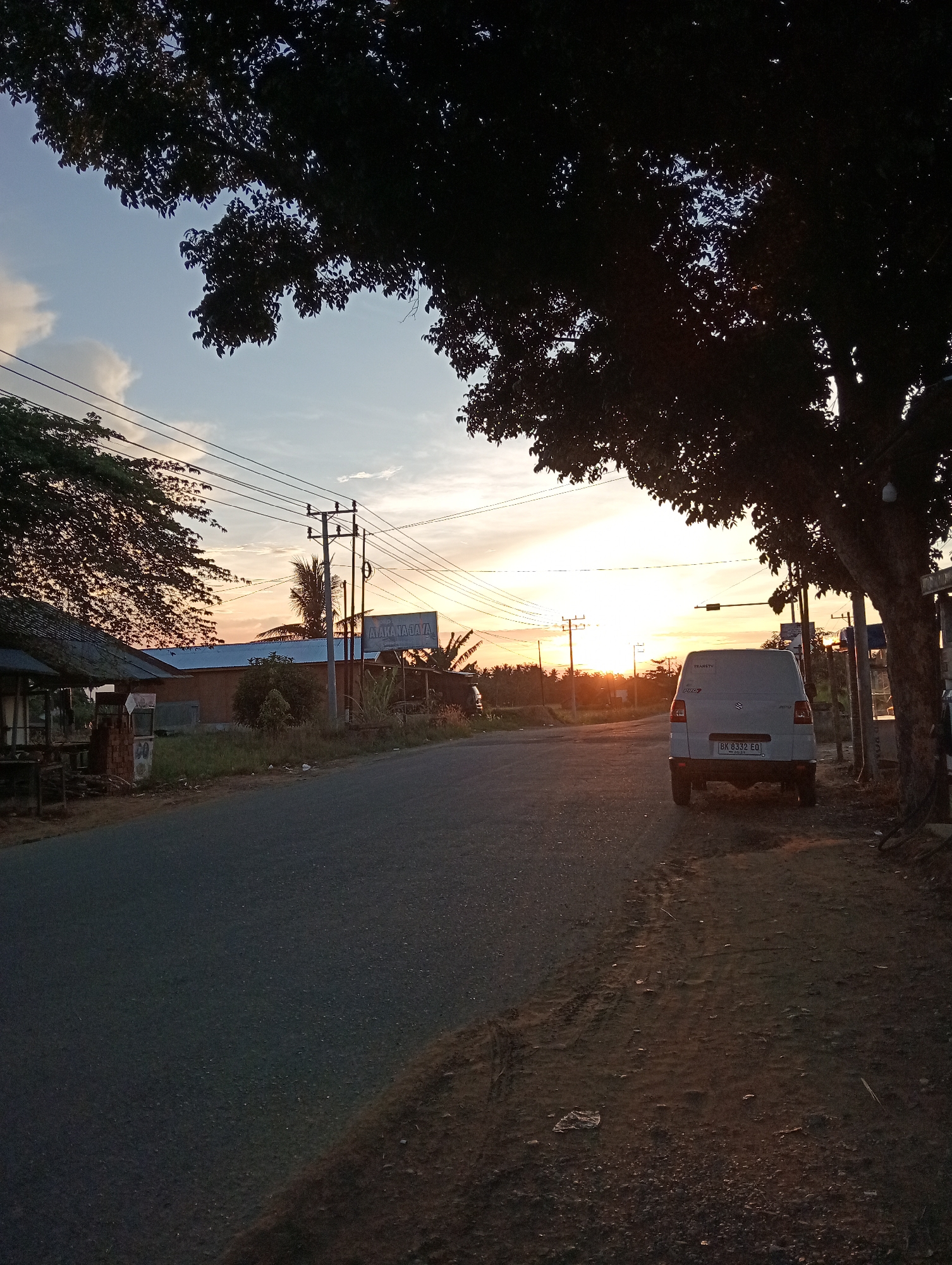 The image depicts a street scene at sunset in an unconfirmed location. A light-colored van is parked on the side of a paved road, near a large tree that partially obscures the setting sun. Power lines run parallel to the road. Across the road, there's a structure with a visible sign "A A A MAJAJA" and a canopy-like structure with a sign above it. The sky shows the colors of sunset. No people are visible.