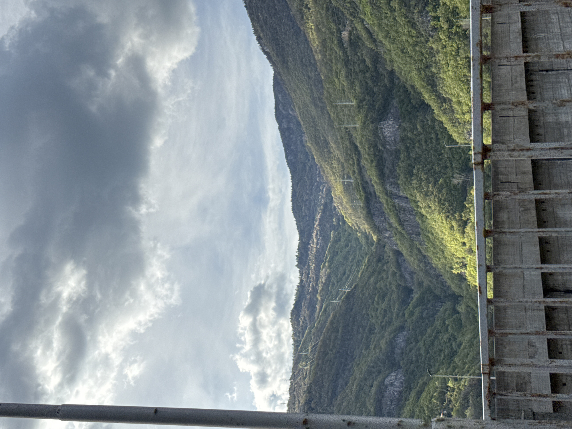The image captures a view in what is likely Mihalkovo, Bulgaria, showcasing a mountain range overgrown with greenery. Power lines traverse the mountain's slope. A weathered concrete structure with regular rectangular cutouts is positioned at the right of the image. A metal rail is in the foreground. The sky is filled with clouds, ranging from lighter to darker hues.