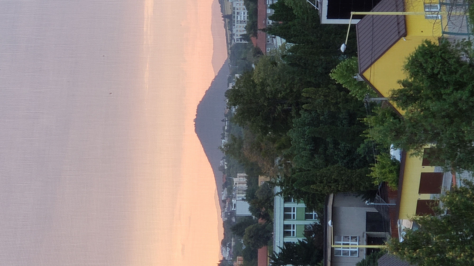 The image, taken from a window with a visible screen mesh, captures a dusky sky with hues of pink and pale blue. In the distance, a prominent, dark, conical mountain dominates the horizon, likely Zemplín Castle Hill (Zemplínsky Hradný Vrch) or a similar prominent peak in the Prešov region, Slovakia. The middle ground features a layered landscape of suburban buildings and dense green trees. Closer to the foreground, the roofs and upper sections of various residential or utility buildings are visible, including a yellow building with a dark brown roof and brown garage doors, and a grey building with a large window. A tall, thin yellow lamppost stands near the yellow building, topped with a white light fixture. The overall scene suggests a view from an elevated position overlooking a semi-urban or suburban area at dusk, with a significant natural landmark in the background.