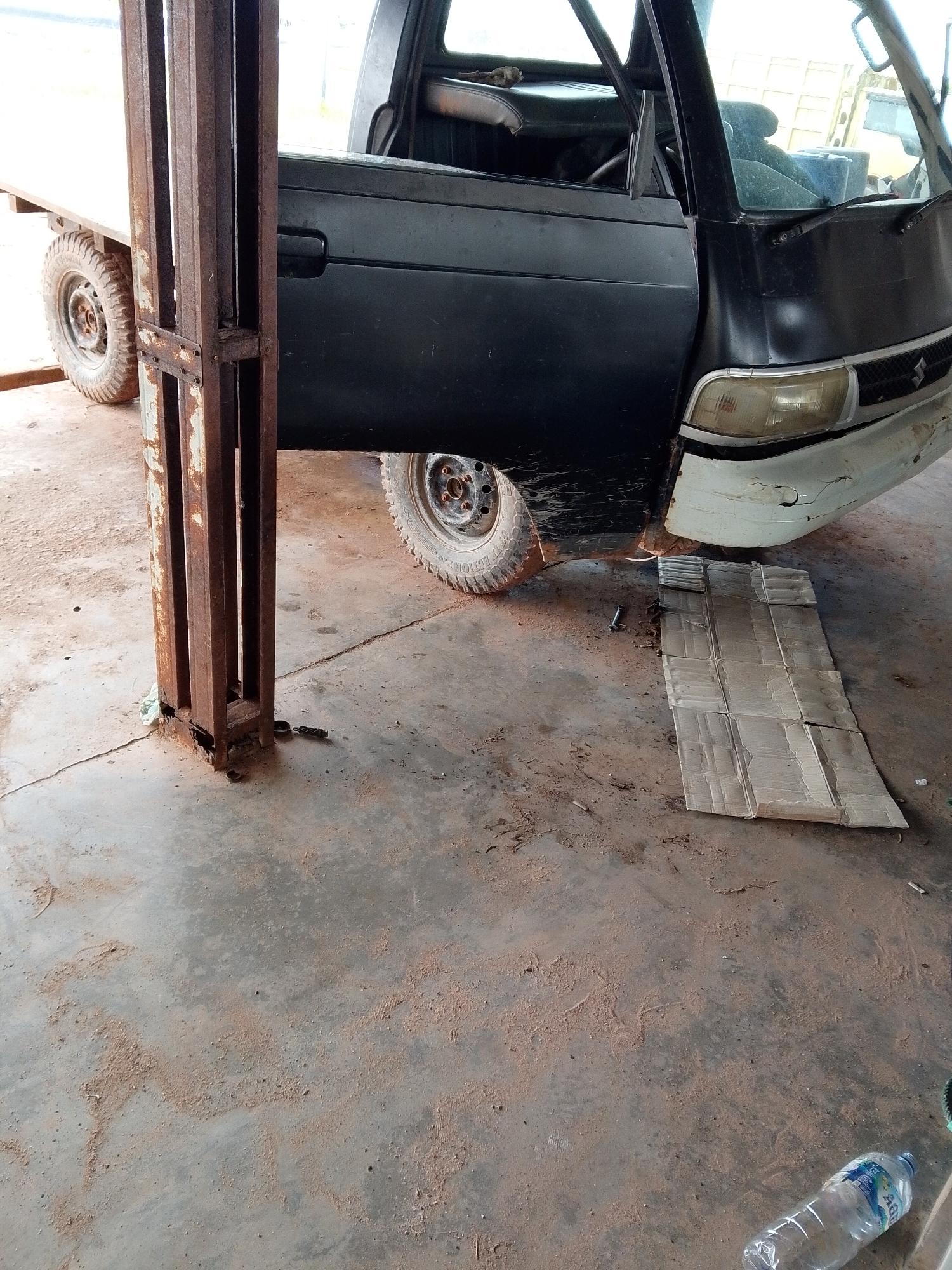 The image shows a damaged black Suzuki Carry pickup truck partially inside what appears to be a garage or workshop. The truck's front passenger side is over a spread of cardboard. The front bumper is visibly damaged. To the left of the truck, a rusty metal support structure stands. The floor is concrete covered with a reddish-brown dirt or dust. In the lower right corner, a plastic water bottle rests on the floor. The location is likely Bukit Ketok, Indonesia.
