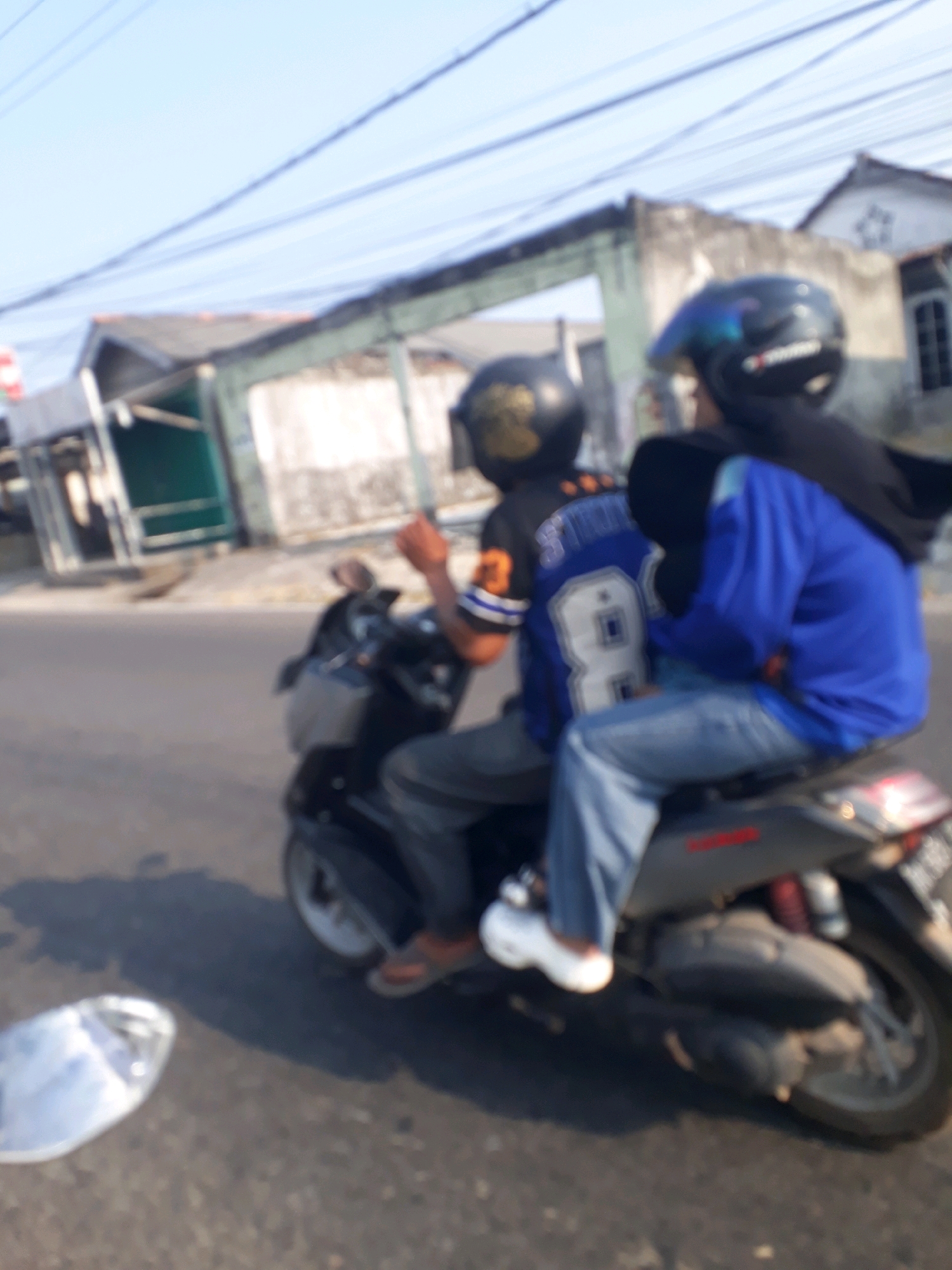 Two individuals are riding on a scooter on a paved road under a bright, clear sky, with telephone wires visible overhead. The person in the front is wearing a dark helmet with blue accents and a dark blue jacket over a black hijab, with light blue jeans. The person in the back wears a dark helmet with gold markings and an orange and black jersey with the number "8" prominently displayed on the back. The scooter appears to be a dark gray model, and the background consists of blurred buildings, possibly shops or houses, with a hint of green structures. The scene suggests a typical day in the city of Pangkalpinang, Indonesia, where scooters are a common mode of transportation. The lighting indicates it is likely daytime, possibly morning or afternoon.