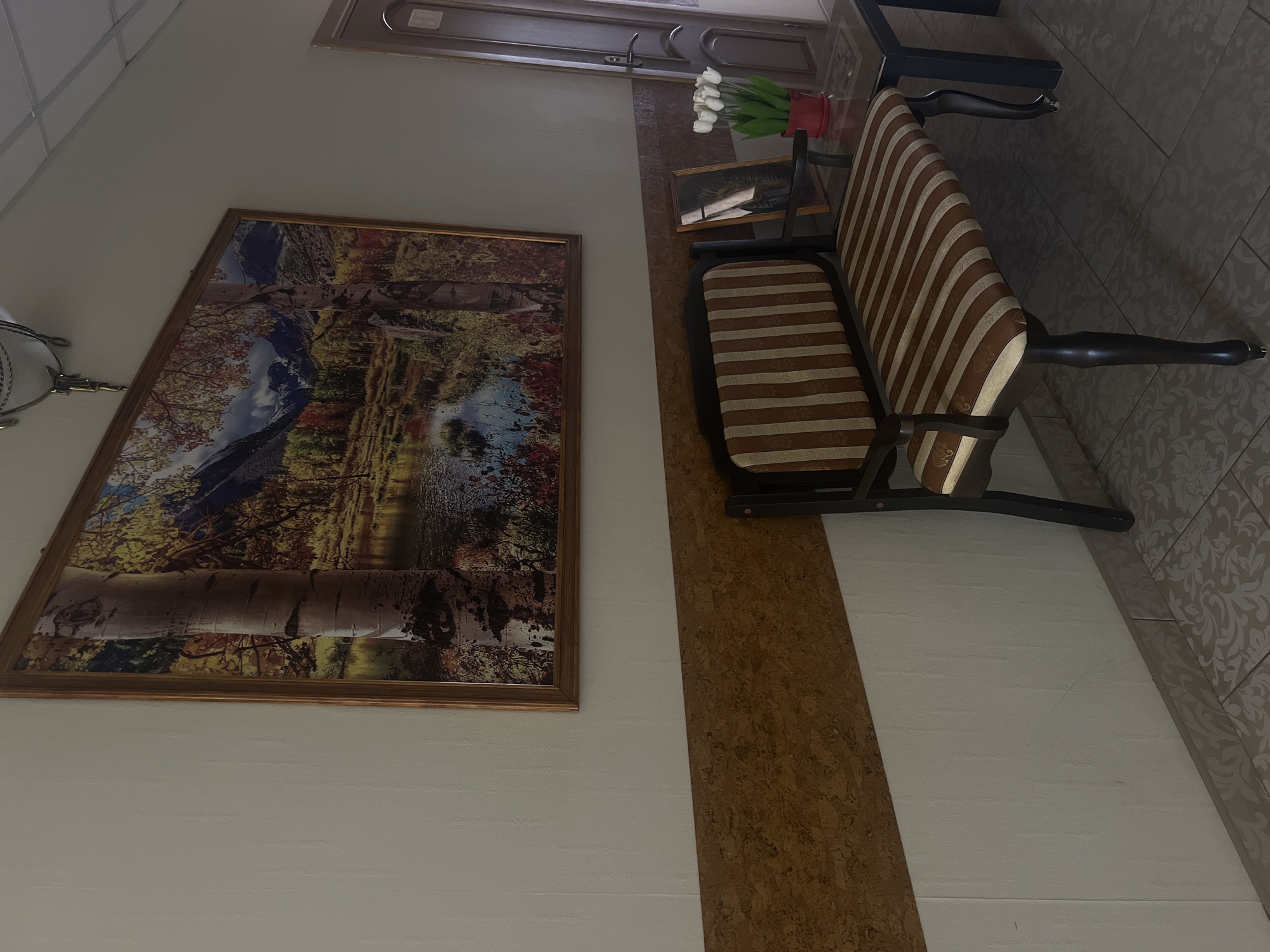 The image shows an indoor hallway, likely in a residential building or hotel, located in Chaikovsky, Russia. A large framed landscape painting depicting a forest scene with a lake and mountains hangs on the wall. Below the painting, a striped upholstered bench sits in front of a wood-paneled section of the wall. To the right of the bench, a small dark table holds a framed photograph and a red pot with white flowers. A door with a dark wood frame and a silver handle is visible on the far right of the image. The floor is tiled with a patterned design. The lighting is somewhat dim, suggesting it might be an interior space without direct sunlight. There are no people visible in the image.