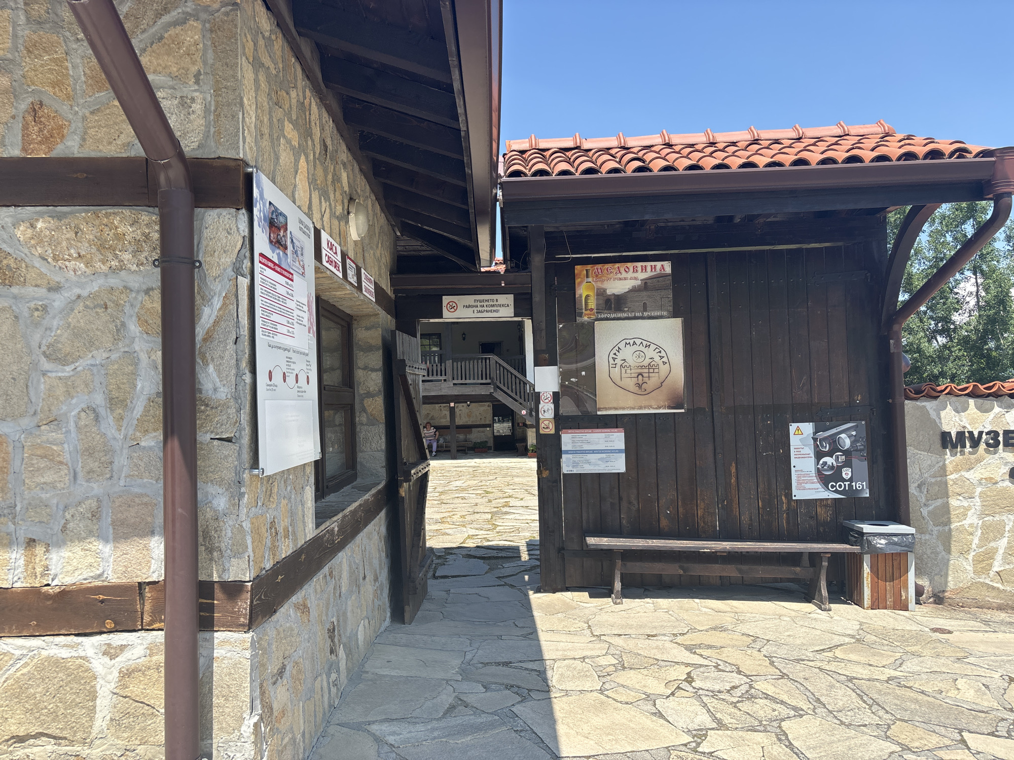 The image depicts the entrance area of a historical or cultural complex in Belchin, Bulgaria, specifically identifiable as part of Tsari Mali Grad due to visible signage. The scene is illuminated by bright sunlight under a clear blue sky.
To the left, a building constructed from irregular, light-colored stone blocks with thick, light mortar features a brown downspout running vertically along its corner. Dark wooden beams are integrated into the stone facade. A white informational sign, detailing operating hours and entrance fees in both Bulgarian and English, is affixed to the wall. Above this sign, partially visible red-on-white signs indicate "KACA" (Cash Desk) and "SOUVENIRS". A window with dark framing is present below the main sign.
In the center, a stone-paved pathway leads into an open passageway between the two main structures. Through this passage, a multi-story building with wooden balconies and stairs is visible in the background. A "No Smoking" sign in Bulgarian ("ПУШЕНЕТО В РАЙОНА НА КОМПЛЕКСА Е ЗАБРАНЕНО!") is prominently displayed within the passage. One individual, possibly a woman in a striped shirt, is seated on a bench in the far background.
To the right, a dark wooden structure, possibly a kiosk or information point, stands with its facade made of vertical wooden planks. It is topped with a terracotta tiled roof and a brown gutter system. Several posters are attached to its wooden wall: one advertises "МЕДОВИНА" (Mead) with an image of a bottle, another features a crest and text likely referring to "ЦАРИ МАЛИ ГРАД", and a third is a warning sign displaying security cameras and the code "COT 161". A long, dark wooden bench is placed in front of this structure, with a dark grey trash can, featuring a wooden base and a black plastic liner, positioned next to its right end. Beyond this structure, another stone wall is visible, with the Bulgarian word "МУЗЕЙ" (Museum) partially carved or affixed to it.
The ground throughout the foreground is paved with irregularly shaped, flat stone slabs. Green foliage from trees is visible in the upper right background, above the roofline.