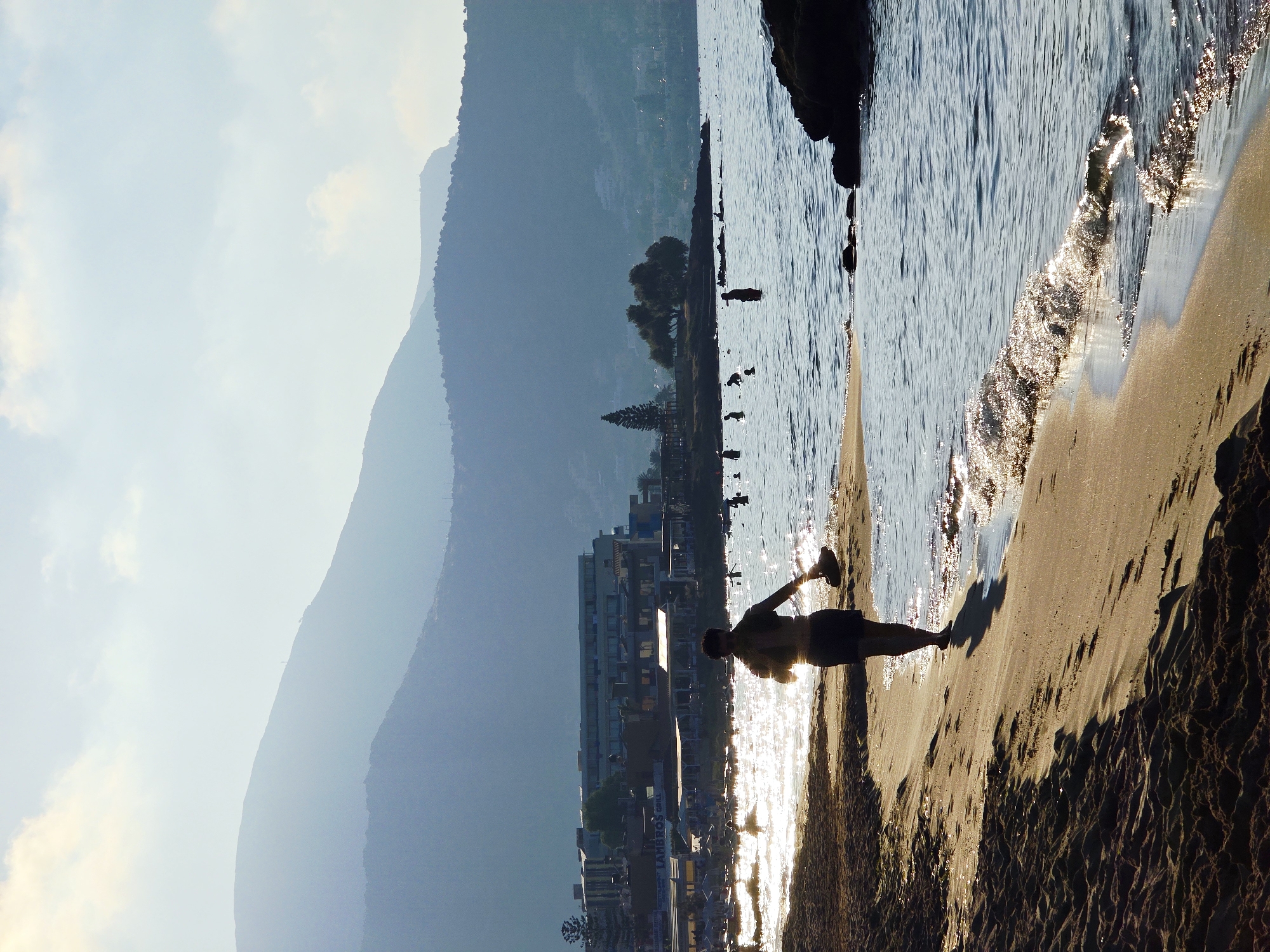 This is a photograph taken at a beach in Malia Municipal Unit, Greece, during the late afternoon or early morning, indicated by the low-angle sunlight creating long shadows and a shimmering effect on the water. The foreground is dominated by a sandy beach, with the waves of the sea gently lapping at the shore. In the mid-ground, a person is walking along the beach, silhouetted against the bright water and sky, carrying a pair of footwear in their hand. The sea is populated with several other individuals, some of whom are wading or swimming.
Further back, a strip of buildings lines the coast, suggesting a developed resort area. These buildings include hotels or apartment complexes and possibly open-air establishments like restaurants or bars, with some visible umbrellas and seating areas. Beyond the coastal developments, layers of hills and mountains rise in the background, fading into a hazy blue due to atmospheric perspective. The sky above is a pale blue with some soft clouds, indicating fair weather. The overall impression is one of a serene and tranquil beach scene.