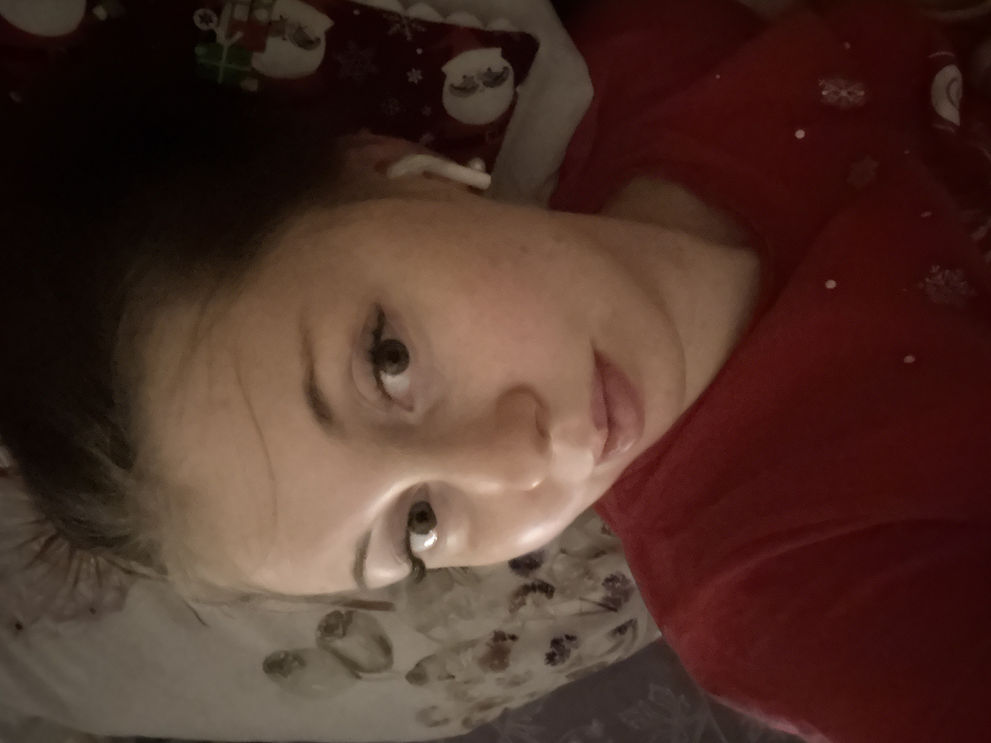 A young person with dark hair and light skin is shown in a close-up, angled shot, appearing to take a selfie while reclining. The person is looking directly towards the camera with a neutral expression. Their eyes are made up with visible eyeliner and mascara. A white earbud is visible in their right ear.
They are wearing a red garment, possibly a shirt or pajamas, adorned with small white snowflake-like patterns. Behind their head is a pillow with a festive, Christmas-themed pattern featuring white Santa heads, snowflakes, and green gift boxes on a dark red background. Another patterned fabric with a floral or abstract design is visible below their chin and neck area.
The lighting in the image is dim and soft, suggesting an indoor setting during evening or night. No specific location cues are visible beyond what appears to be a bed or couch. There is no visible text in the image.