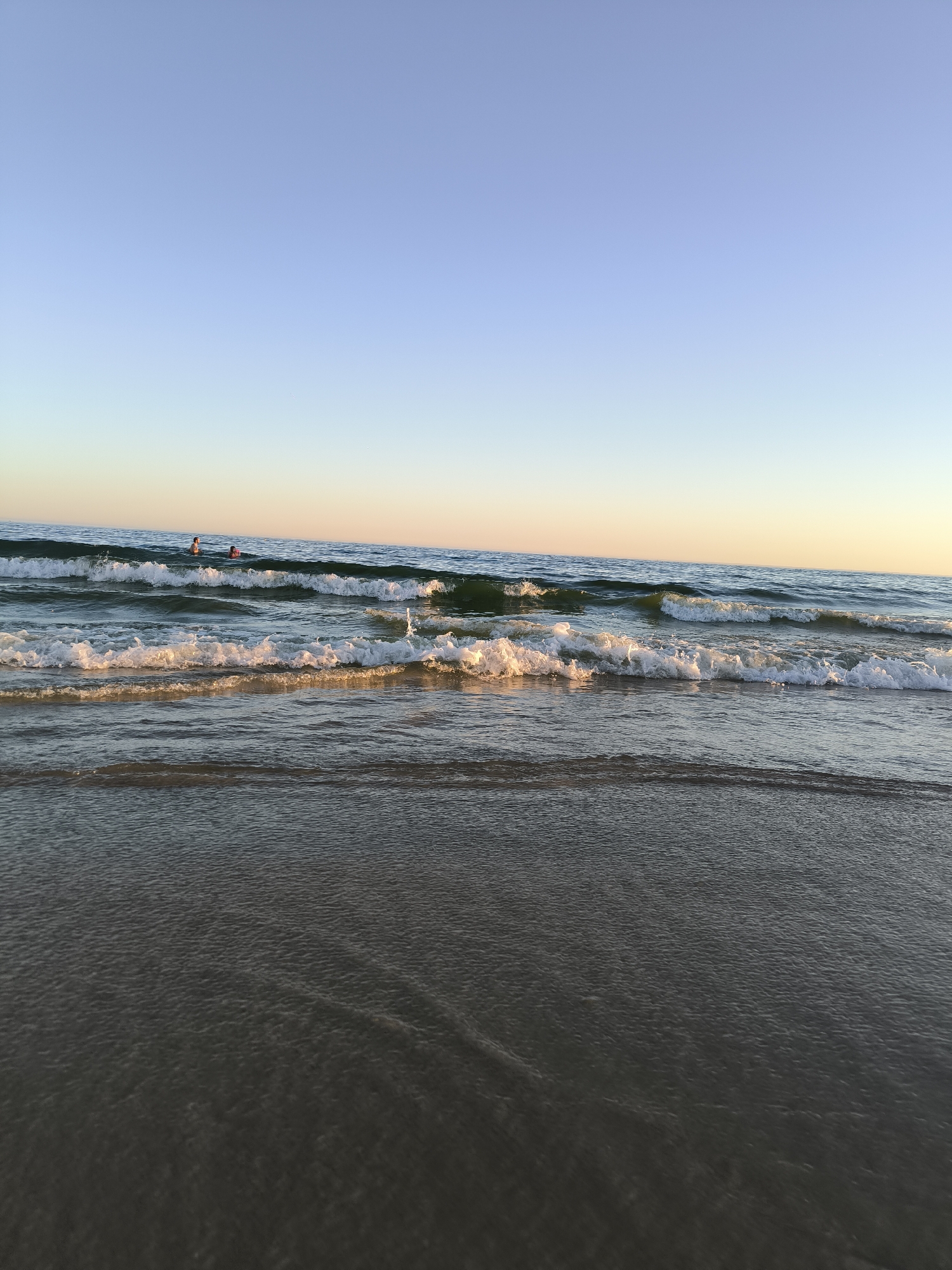 This is a landscape photograph taken at sunset on the beach of Vila Real de Santo António, Portugal. The image captures the sea with gentle waves rolling towards the shore, and two individuals are visible swimming in the distance. The sky is a clear gradient of blue, transitioning to a warm, pale yellow at the horizon. The foreground is dominated by wet sand, reflecting the soft light of the setting sun. The overall scene is serene and tranquil.