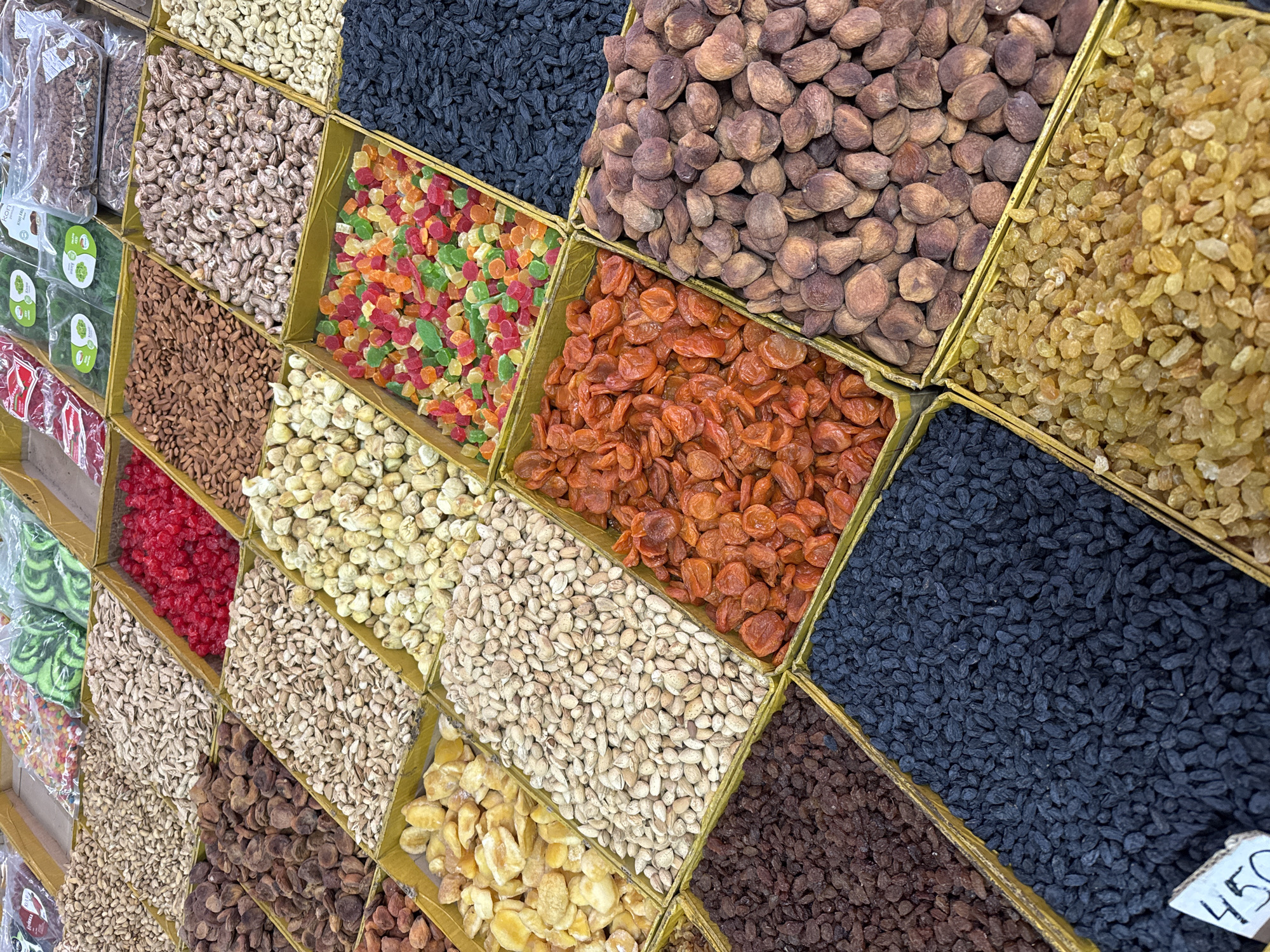 A vibrant display of dried fruits, nuts, and candies is presented for sale in a market stall, likely in Bishkek City, Kyrgyzstan. The items are neatly arranged in numerous rectangular and square bins, forming a colorful grid viewed from a slightly elevated perspective.
The scene showcases a rich variety of goods. Visible items include multiple types of cashews (some light-colored, others with darker skins), dark brown dried apricots, bright orange dried apricots, golden raisins, and dark black raisins (possibly currants). Candied fruits in cubes of red, green, orange, and yellow hues add splashes of color. Various nuts are also present, such as almonds (both shelled and with skins) and light-colored shelled pistachios. Other dried fruits like red candied cherries or berries, yellow/pale orange chunks (possibly pineapple or pears), and different shades of brown raisins complete the assortment.
On the far left, some items are visible in clear plastic packaging, including green leaf-shaped candies and red packets. The bins appear to be simple wooden or cardboard containers, with some possibly lined with a gold-colored material. The lighting is bright and even, typical of an indoor market setting. In the bottom right corner, a white price tag with the number "450" is visible, likely indicating the cost in Kyrgyzstani Som. No people are visible, but the organized display clearly indicates a commercial setup for customers to browse and purchase.