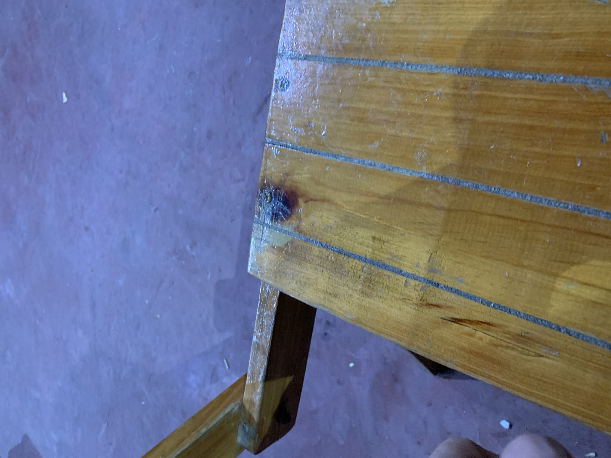 The image presents a close-up, high-angle view of a worn wooden surface, likely part of outdoor furniture such as a bench or table, or possibly a boat deck, in Santa Ana, Venezuela. The surface is composed of several light-brown wooden planks arranged horizontally, separated by dark gray, textured lines, possibly caulking or sealant. The wood exhibits a glossy, somewhat faded and possibly wet or dirty varnish or finish.
A prominent dark, charred mark is visible on the upper left section of the wooden surface, where faint, capital letters, possibly "CO" or "GO," can be discerned within the damaged area. Further down and to the right, a long, dark scratch or stain mars another plank. A wooden support structure, angled downwards, is partially visible on the left side, extending from beneath the main wooden surface.
The ground beneath the structure is a textured, reddish-purple color, suggestive of concrete or paving stones, with small white specks scattered across it. In the very bottom right corner of the image, the toes of a human foot are visible, indicating the presence of a person near the object. The lighting appears subdued, making the specific time of day unclear. There is no explicit activity taking place; the scene focuses on the physical state of the wooden object and its immediate surroundings.