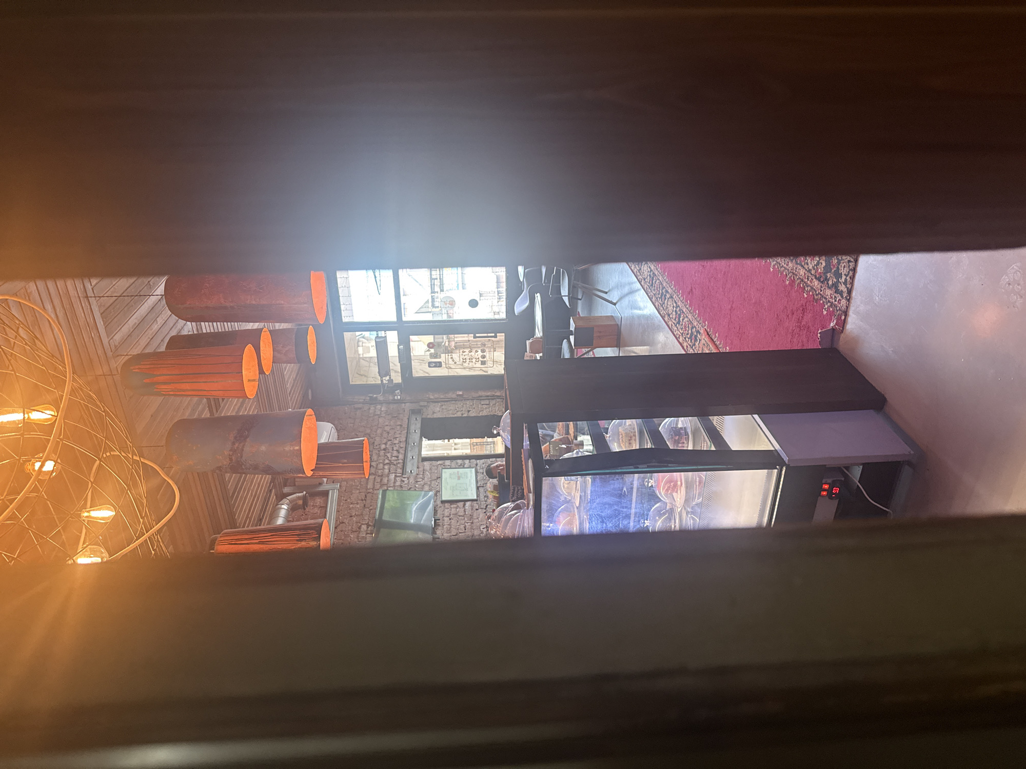 The image depicts the interior of a cafe, viewed from a doorway. The cafe has a rustic ambiance with exposed brick walls and a wooden ceiling. Several large, cylindrical lamps with orange interiors hang from the ceiling, casting a warm glow. A glass display case showcasing cakes and pastries is prominently featured on the left side of the frame. Behind the display case, a person is visible behind a counter, interacting with a customer or preparing items. Tables and chairs are arranged in the background, some near a window that allows natural light to enter. A red carpet with an ornate pattern runs across a portion of the floor, adding a touch of color to the scene. The overall impression is of a cozy and inviting establishment. It is not possible to determine the specific location in Birobidzhan, Russia from the image alone.