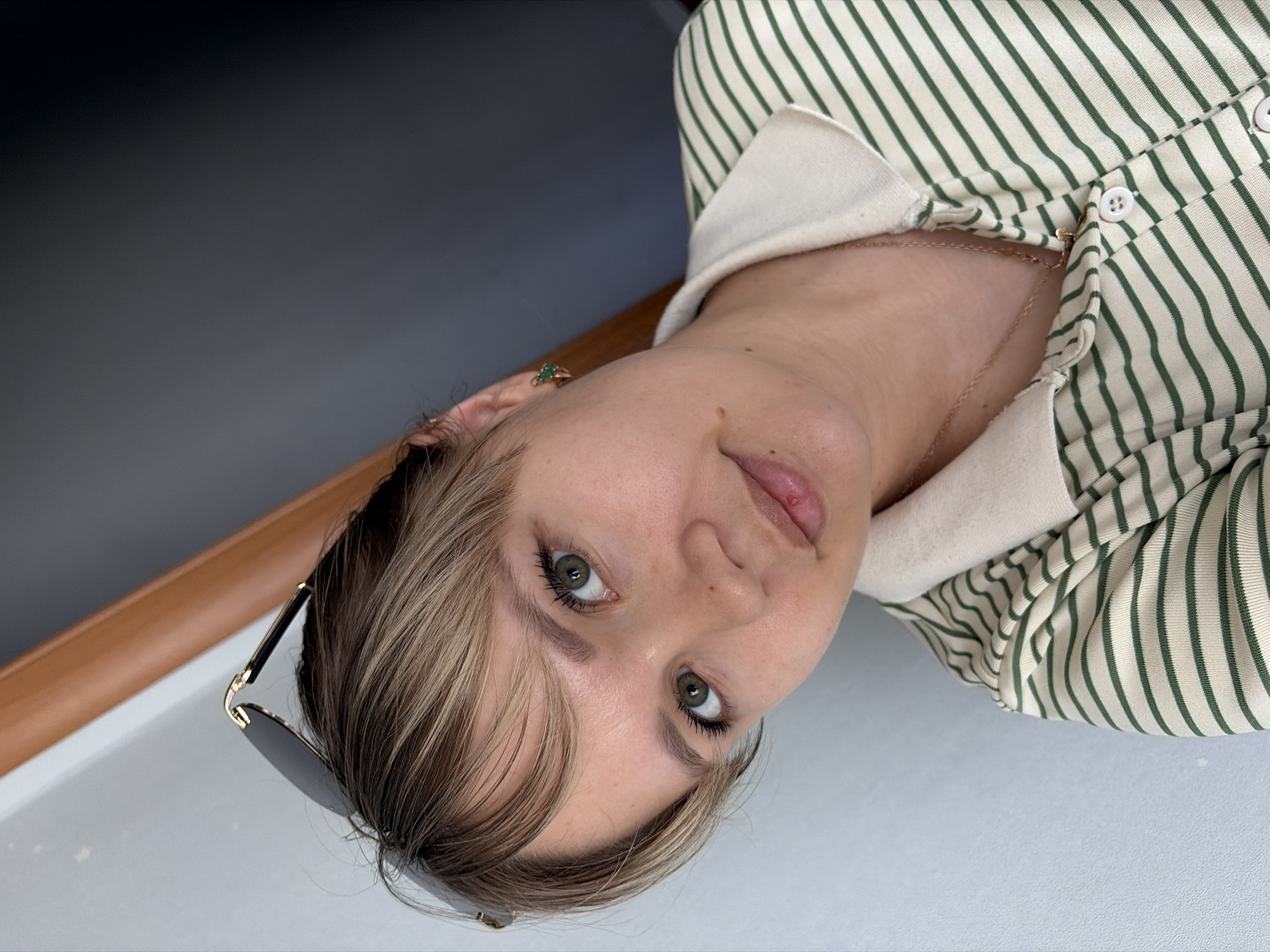 A close-up, slightly off-center portrait shows a woman with light skin and blonde, highlighted hair. Sunglasses are perched on her head. She has green eyes and appears to be looking directly at the camera. She is wearing a button-down shirt with green stripes and a white collar. A delicate gold necklace is visible around her neck, and she has a green stud earring in her left ear. The background includes a portion of a wooden railing and a grey or black surface, likely a wall or table.
There is no information to suggest the location is Chaikovsky, Russia.