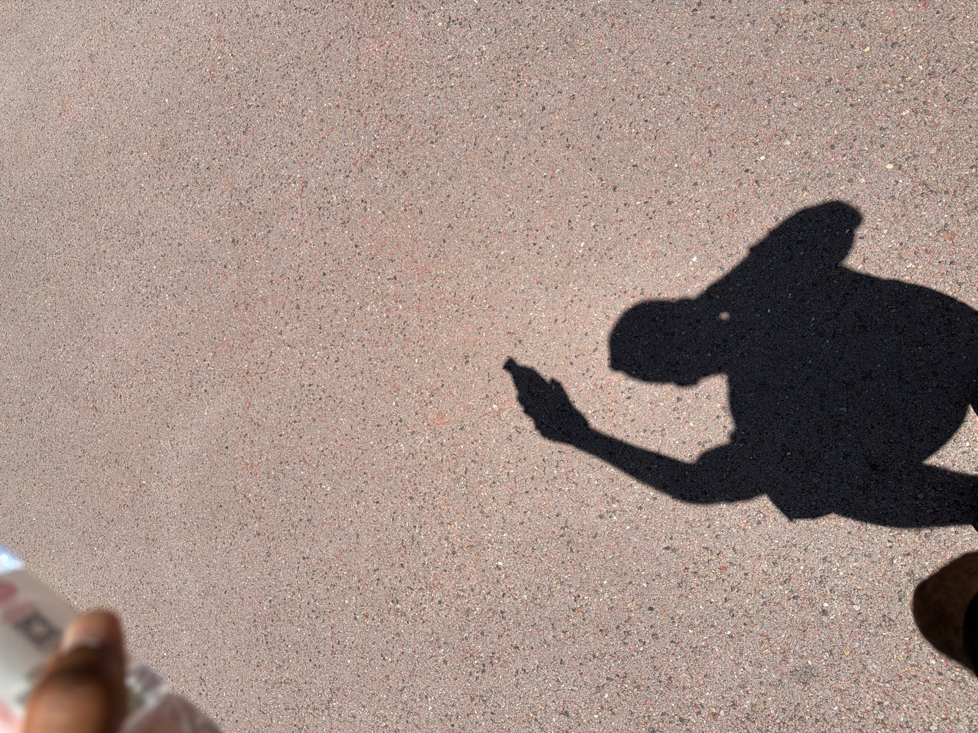 A shadow of a person is cast on a light brown, coarse-textured surface, possibly asphalt or concrete. The person appears to be standing, with one arm raised and bent at the elbow, as if shielding their eyes from the sun. A portion of a hand holding a white object, possibly a bottle or container, is visible in the upper left corner of the image, casting its own faint shadow. The environment is outdoors under bright sunlight. The image does not contain information to determine the specific location within Jiangjin, China.