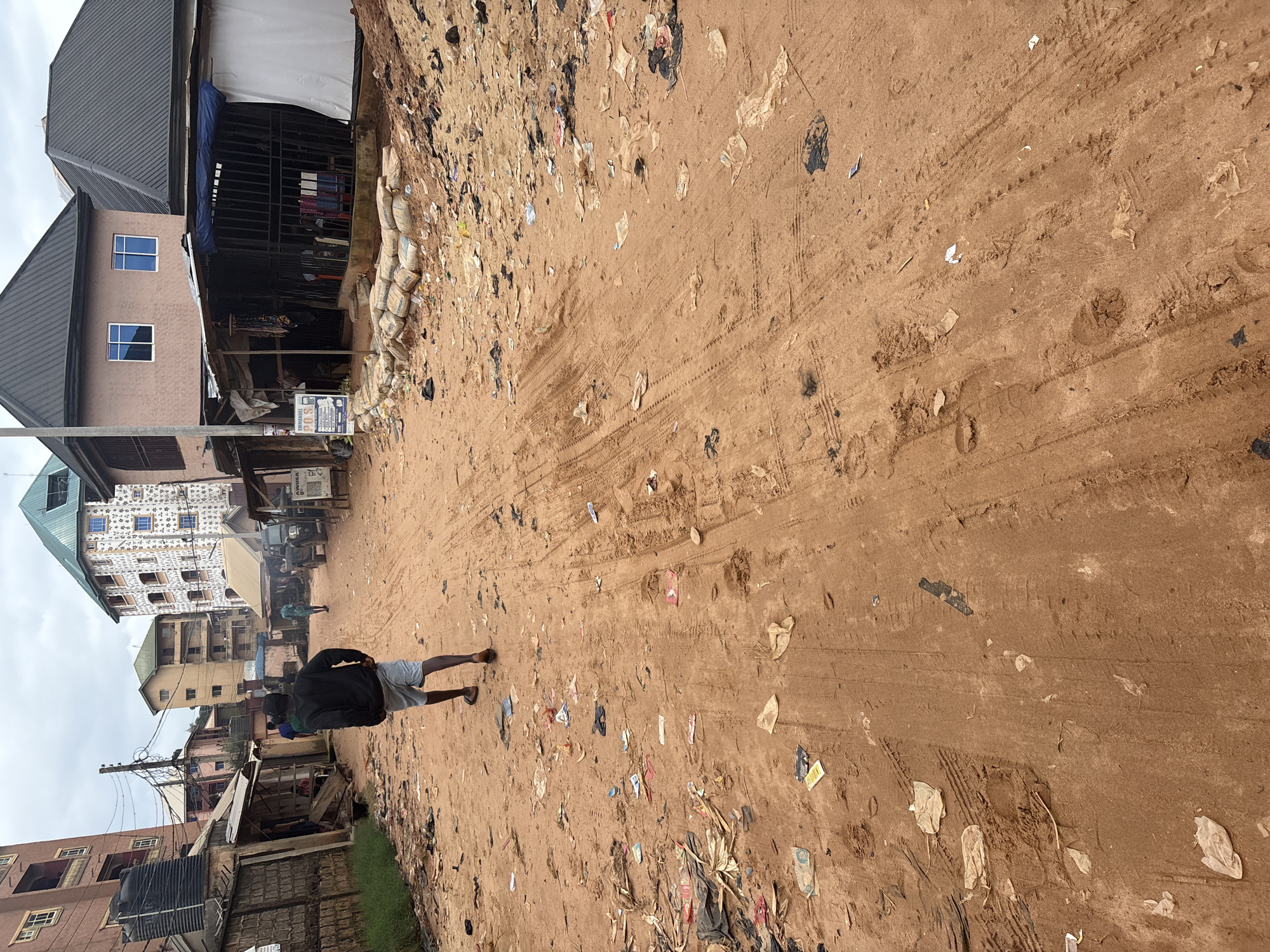 The image depicts a street scene in Nkpor, Nigeria. A person, wearing a black jacket and grey shorts, walks away from the camera down a dirt road. The road is uneven and appears to be a dirt track with tire marks and footprints. It is littered with various types of debris, including plastic bags, wrappers, and other unidentifiable trash. On either side of the road, there are buildings. To the left, there are multi-story buildings with balconies and what appears to be a water tank. There are also structures resembling market stalls with corrugated metal roofs. To the right, more buildings are visible, including a multi-story structure with a green, pointed roof and a building with a pink facade and dark grey pitched roofs. In front of one of the buildings on the right, there are sacks of what appears to be cement piled up. Several other people are visible in the background, some walking and others standing near the stalls. The sky is overcast.