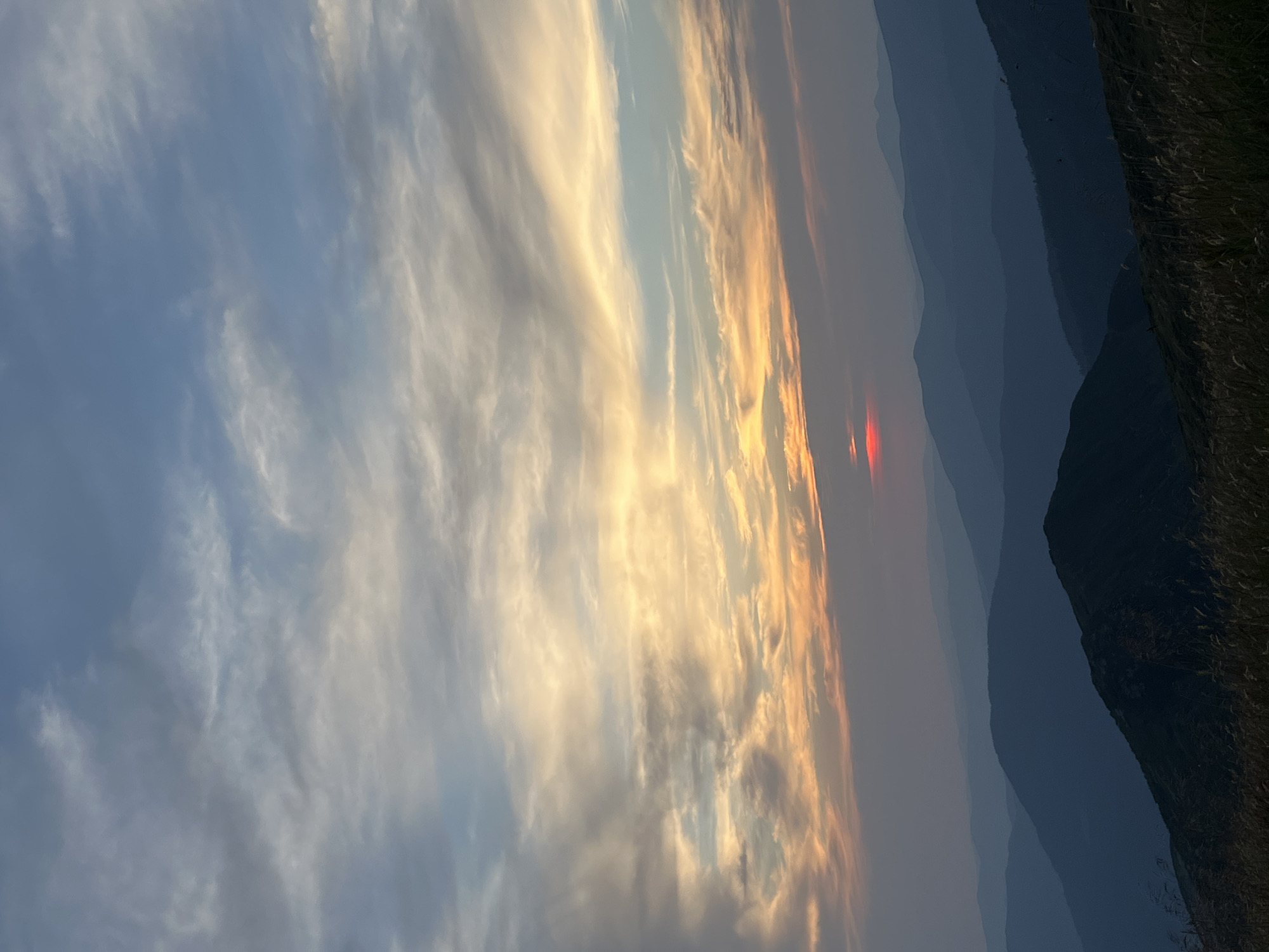 The image depicts a panoramic view of layered mountain ranges under a vast sky, likely taken at dusk. The sky is filled with wispy clouds, painted with hues of orange, yellow, and blue, indicating the presence of a setting sun. A small, vivid red glow, likely the sun itself, is visible on the horizon, nestled between the upper ridges of the mountains. The mountains are depicted in shades of deep blue and purple, with each successive range fading into the atmospheric haze, creating a sense of depth and distance. In the foreground, the silhouette of a dark, grassy slope is visible, with dry grass stalks adding texture. The overall scene suggests a remote, natural landscape. The location is identified as the city of Luhy, Ukraine. There are no people or animals visible in the image, nor any man-made structures or objects, indicating a focus on the natural beauty of the mountainous terrain and the sky.