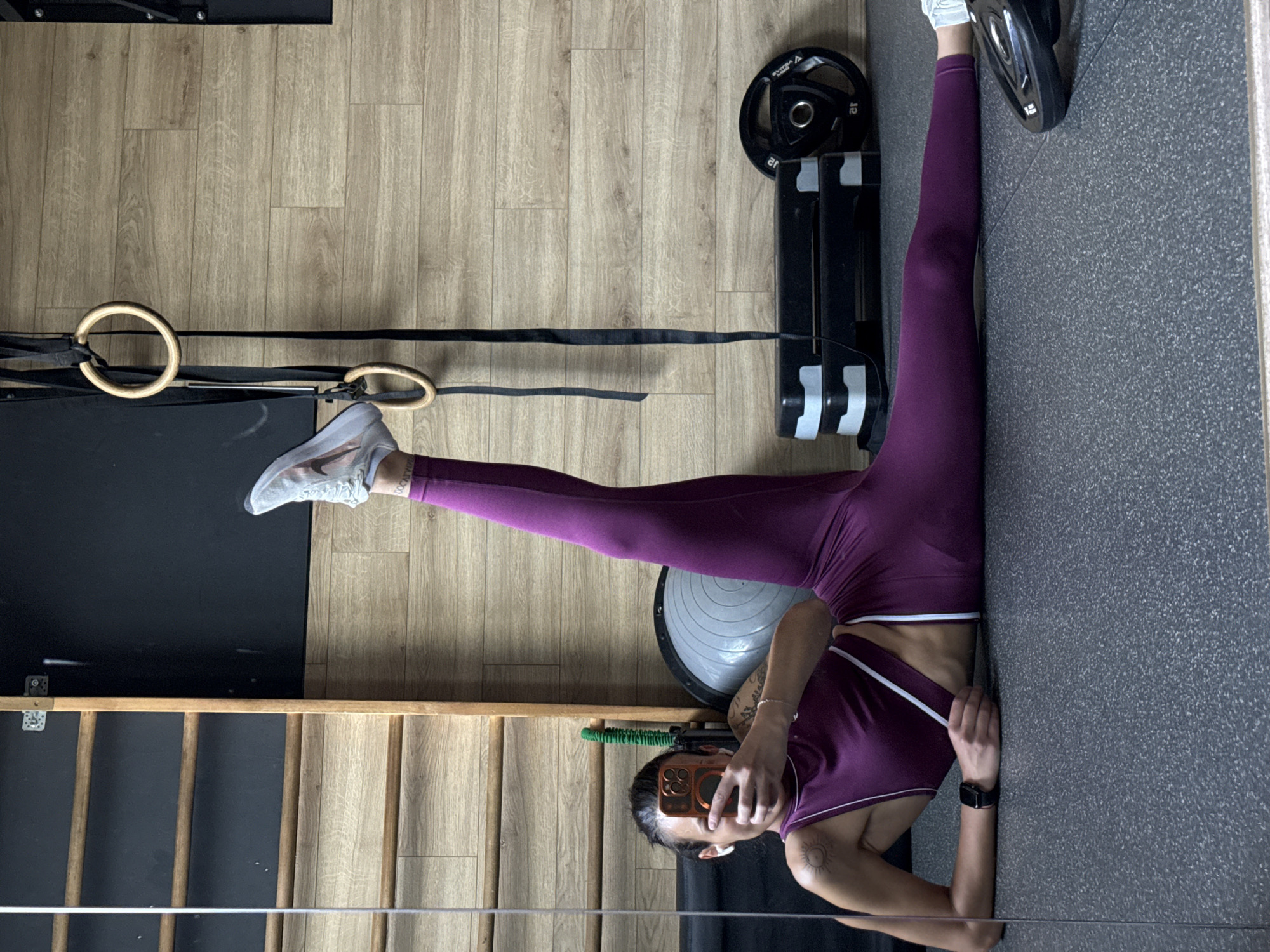 A person in workout clothes and shoes is taking a selfie in a room that appears to be a gym or workout space. The person is positioned with their back against a textured gray wall, legs extended upward in a split position. Gym equipment is visible in the background. The location cannot be determined to be in the city of Pleven, Bulgaria.