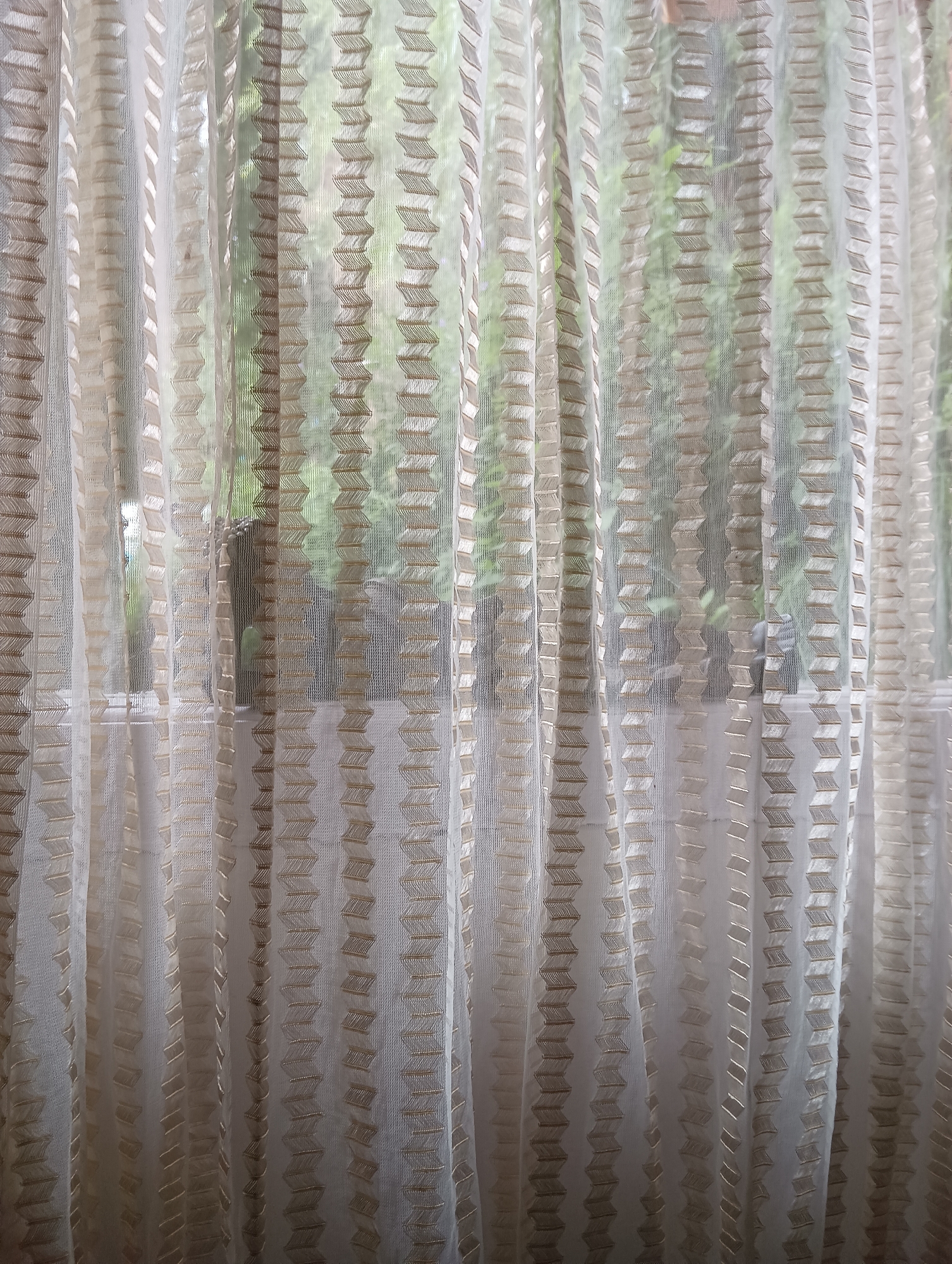 A close-up shot of sheer, white curtains with a decorative pattern of golden, zig-zagging lines running vertically. The curtains are layered and draped, creating a soft, textured effect. Through the translucent fabric, the blurry shapes of green foliage and a hint of what appears to be a window frame are visible, suggesting an indoor setting looking out onto an outdoor environment. The lighting is soft, with some areas of the curtain appearing brighter due to light filtering through. The overall impression is one of gentle diffusion and subtle detail.