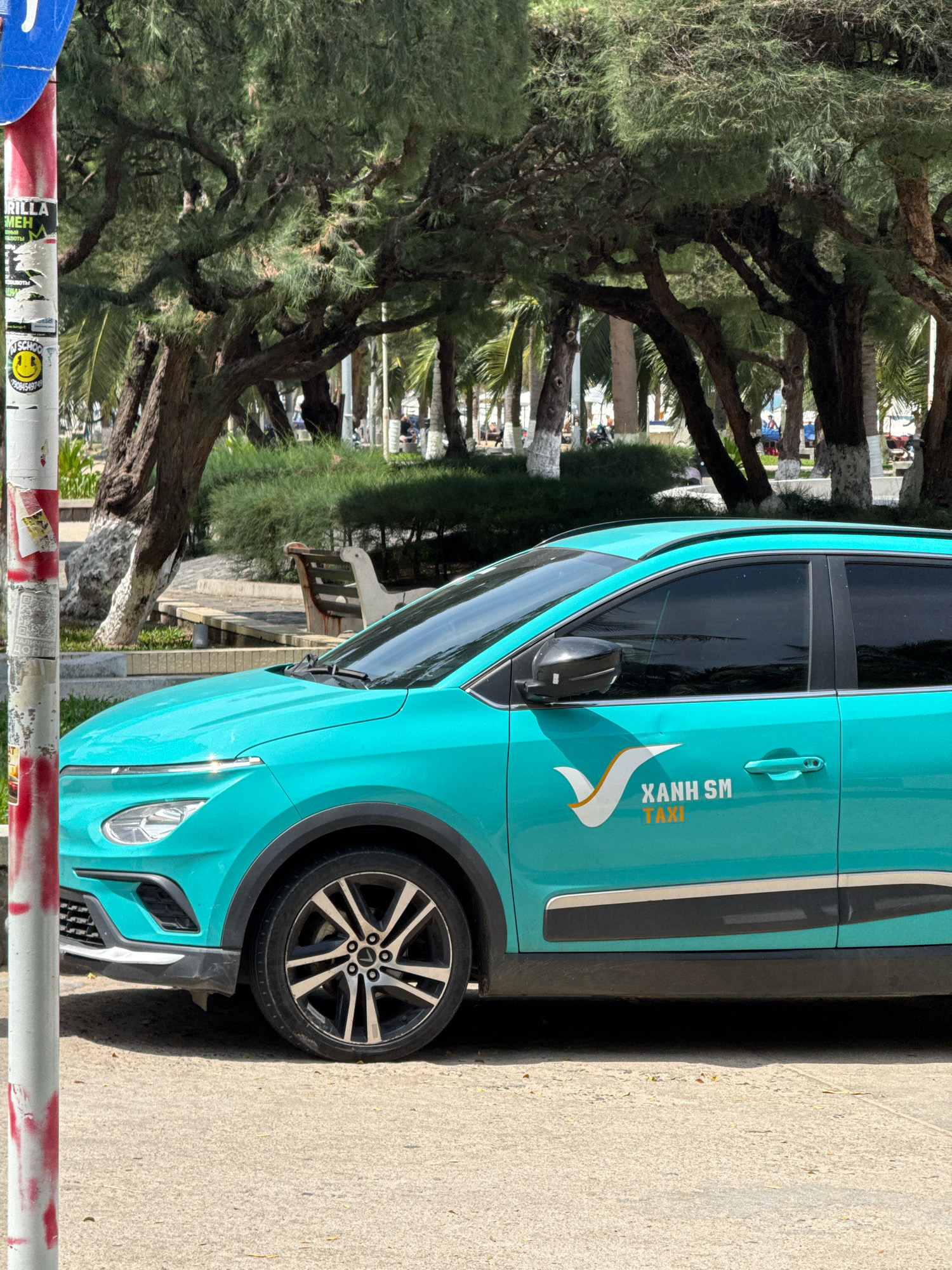 A bright turquoise taxi, identified by the "XANH SM TAXI" logo, is parked on a paved surface. The taxi is a modern compact SUV with tinted windows and black alloy wheels. In the background, a park area is visible with mature trees, palm trees, and a white bench. A red and white striped pole with various stickers is in the foreground to the left of the taxi. The scene appears to be in a public outdoor space, likely a street or parking area adjacent to a park, consistent with a location in Nha Trang, Vietnam, given the taxi branding. No people are prominently featured in the image.
