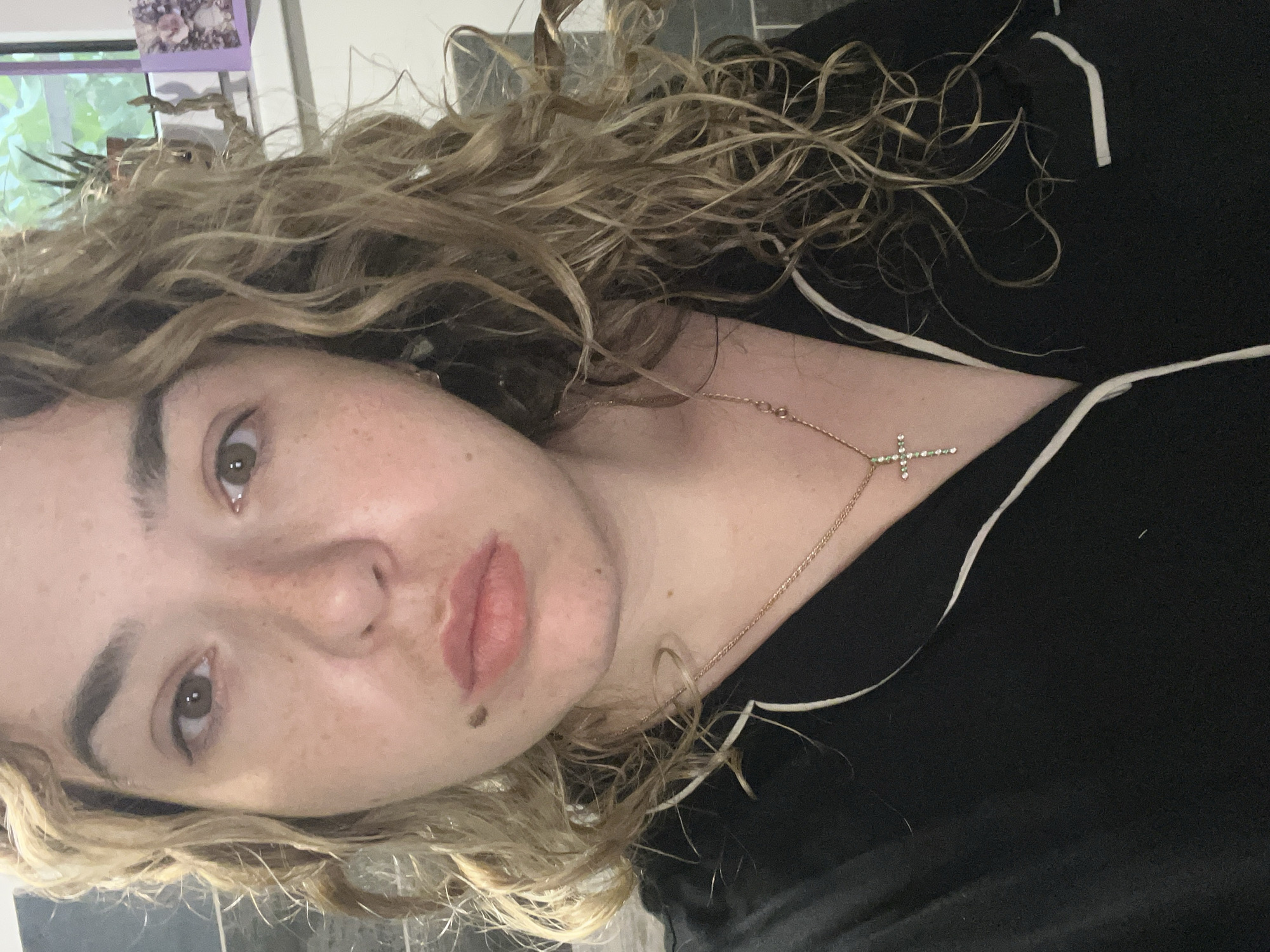 The image is a close-up selfie of a woman with curly blonde hair. She is wearing a black pajama top with white trim and a gold necklace with a jeweled cross pendant. She has freckles on her face and a mole on her left cheek. The background is blurred, but appears to be indoors with some plants and a window visible. The location is not specified in the image.
