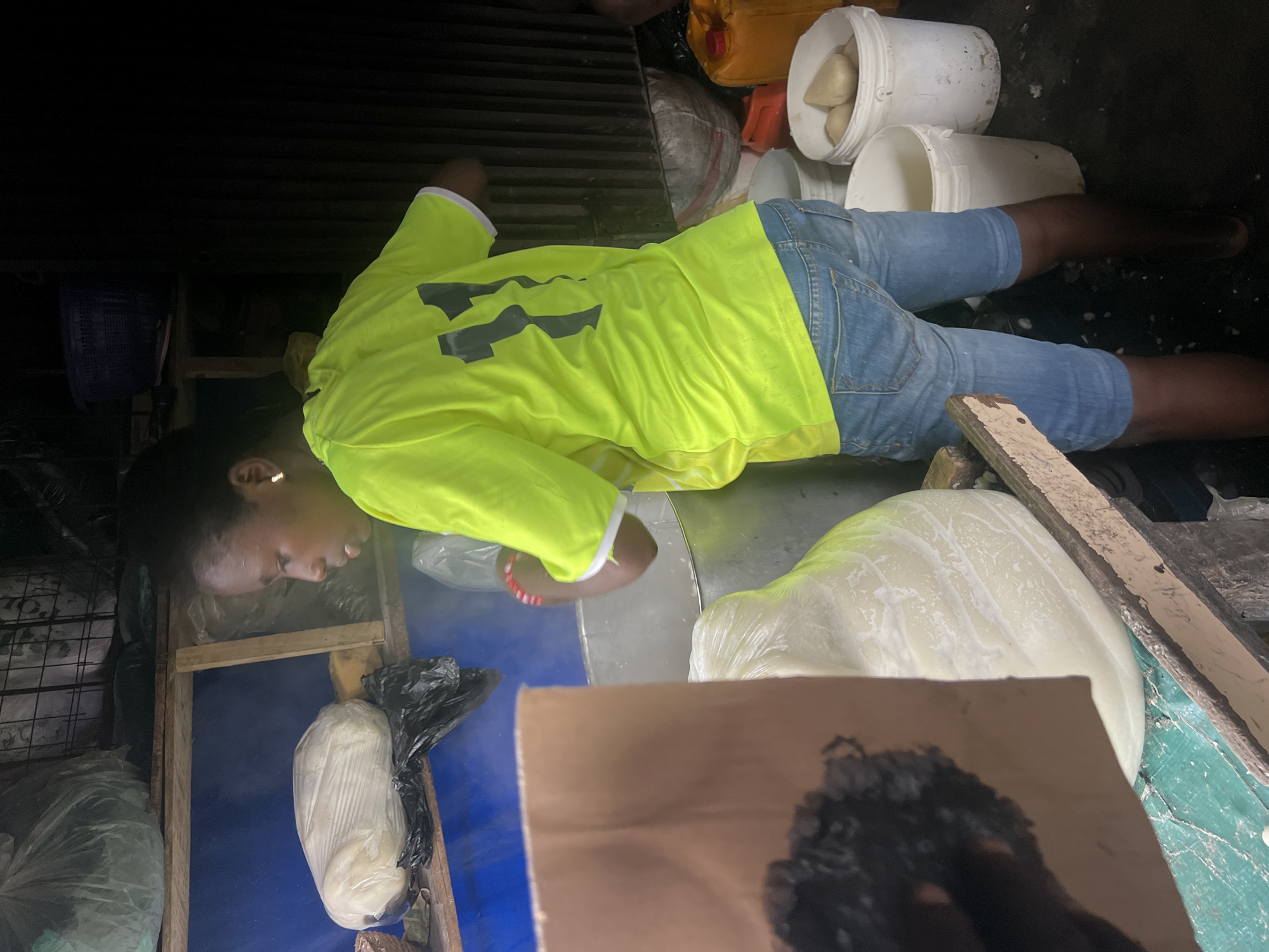 A young woman wearing a neon yellow t-shirt with the number "11" on the back and blue denim shorts is preparing food in an outdoor market stall in Nkpor, Nigeria. Steam rises from a large metal pot in front of her, indicating the food is cooking. A large, white, dough-like mass, possibly yam or cassava, is visible in a plastic bag within the pot. To the left, a large, wrapped bundle of what appears to be food is on a shelf, alongside black plastic bags and a woven basket. In the background, shelves hold more items, and to the right, several white plastic buckets are filled with more food items and a large yellow jerrycan is present. The ground is a mix of dark earth and a green tarp. The scene suggests a food preparation area within a busy marketplace.