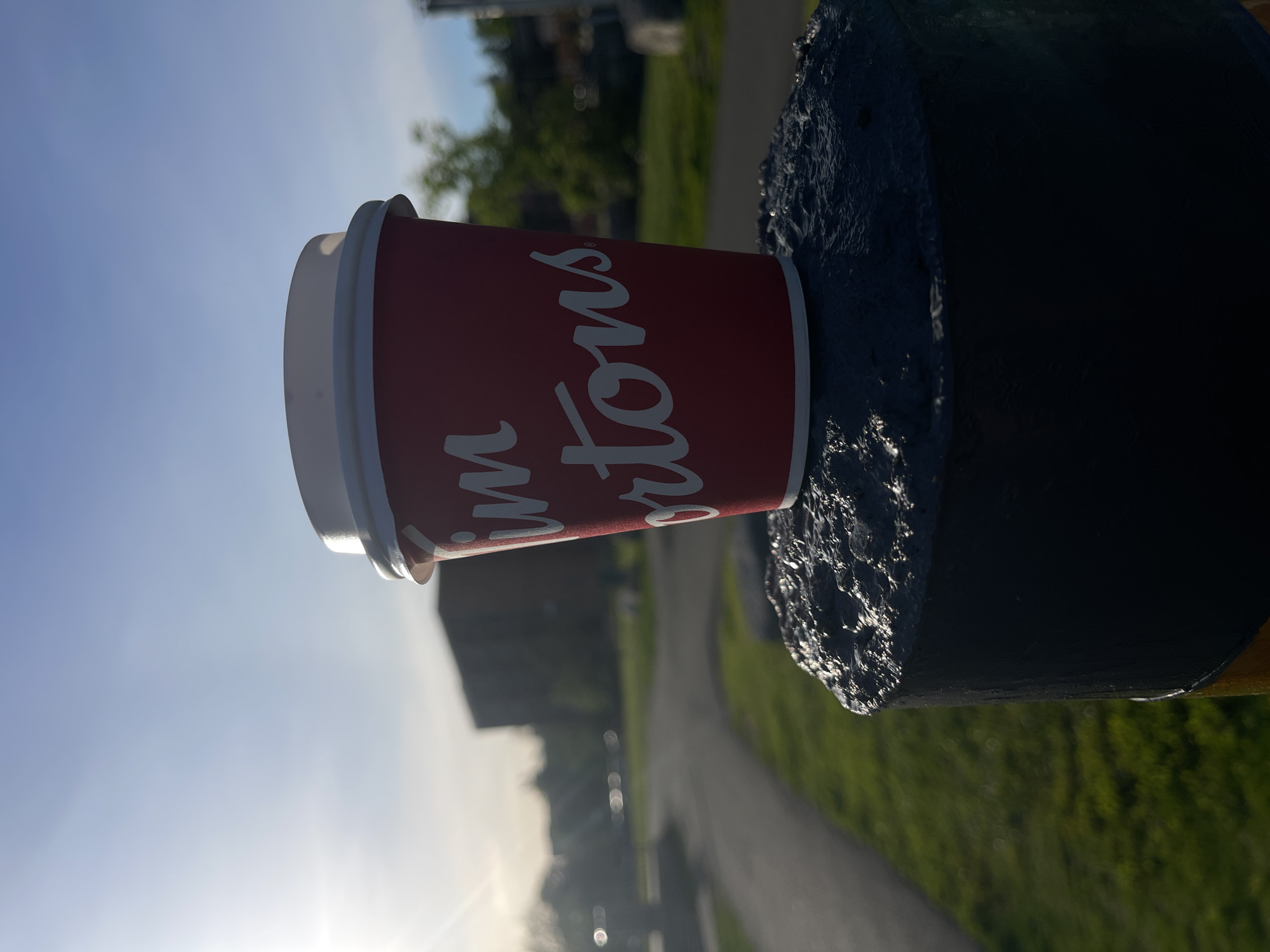 The image depicts a red and white Tim Hortons coffee cup, prominently featuring the brand's white cursive logo on its red body. The cup appears to be standing upright, resting against a dark, textured object, likely a painted pole or bollard, which occupies the right side of the frame. A white plastic lid is visible on top of the cup.
The background reveals a clear blue sky, with the sun low on the horizon, casting a bright glow from the left side of the frame, suggesting either early morning or late afternoon. A paved path, possibly asphalt or concrete, curves into the distance on the right, bordered by vibrant green grass and trees. Blurred buildings or structures are visible in the far background. This outdoor setting, characterized by clear, sunny weather, suggests a park or a pathway in an urban or suburban area, consistent with the provided location context of Ottawa, Canada. No specific activity or individuals are present. The only visible text is "Tim Hortons" on the cup.