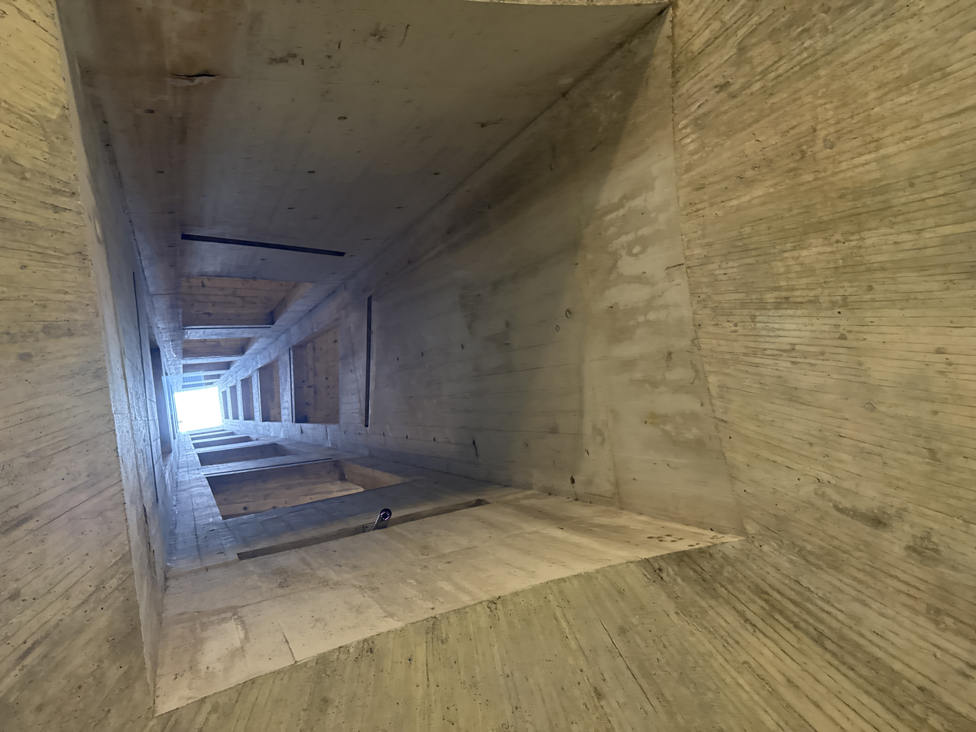 The image depicts the interior of a brutalist concrete structure, viewed from below, looking upwards towards a bright opening. The walls and ceiling are made of raw, textured concrete, exhibiting the marks of wooden formwork used during construction. The space appears to be a vertical shaft, possibly an elevator shaft or a ventilation duct, with a series of openings or windows visible on the far side, arranged in a regular pattern. Light streams in from the top, creating a strong contrast with the dark, shadowed interior. The overall impression is one of stark, industrial architecture. This structure is located at Castel Grande in Bellinzona, Switzerland, a UNESCO World Heritage site, where a modern elevator and walkway have been integrated into the historic fortress.