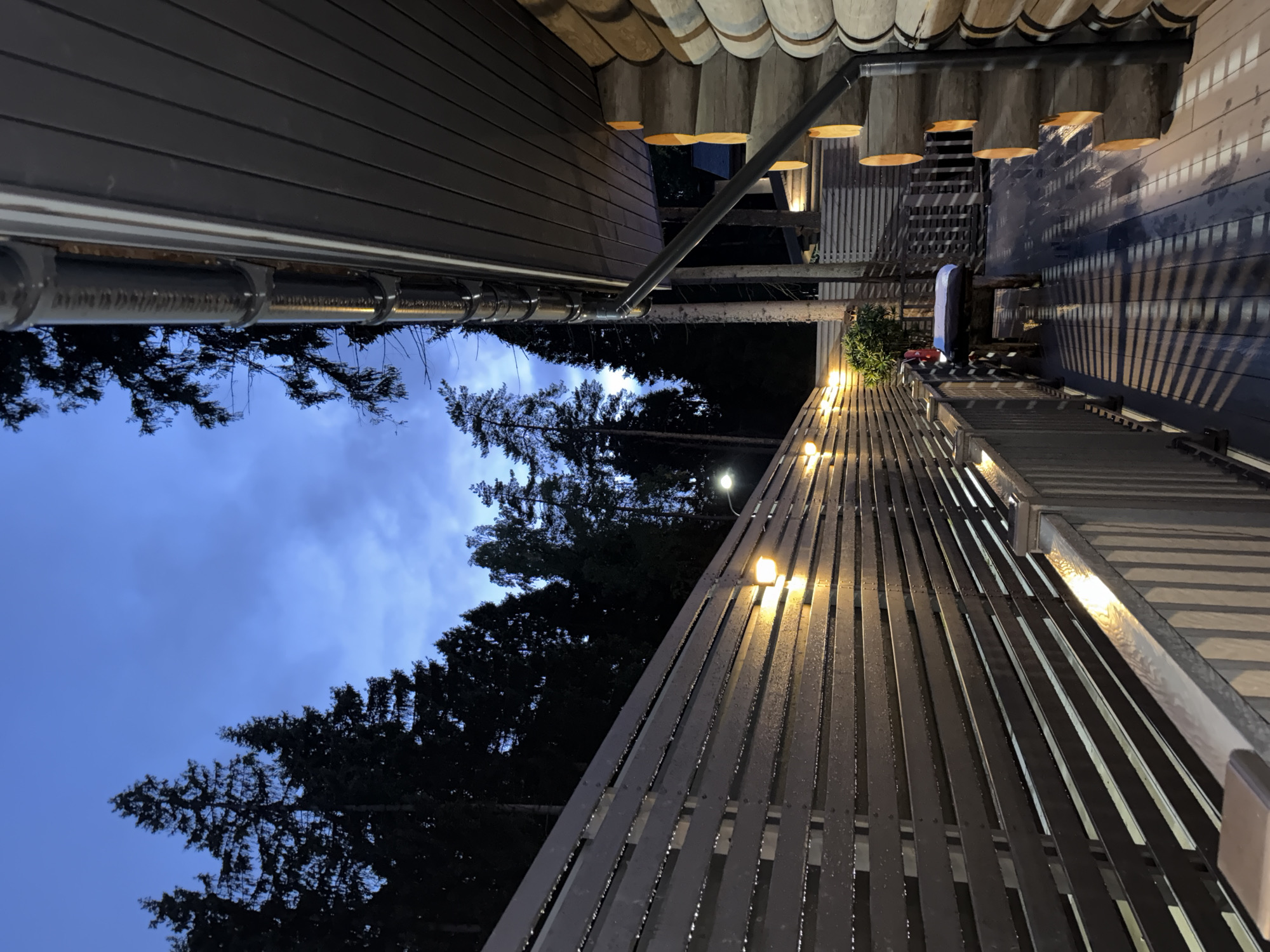 The image depicts an outdoor deck area at dusk, illuminated by artificial lights. On the right, a log cabin structure is visible, with its rounded wooden walls stacked horizontally. A dark-colored downspout runs vertically along the side of the cabin.
To the left of the cabin, a wooden deck extends outwards. A horizontal wooden fence, also lit by embedded lights, runs along the edge of the deck. The fence appears to be made of horizontal slats. The deck itself is a dark wood surface with diagonal shadows cast by the fence and overhead structures.
In the background, a dense forest of tall, dark coniferous trees dominates the landscape under a twilight sky with hints of blue and gray clouds. One visible street lamp is positioned near the fence, further illuminating the scene. There is a small, dark-colored wooden bench with a cushion on the deck. No people are visible in the image. The scene appears to be located in a rural or wooded area, potentially in the vicinity of Novyy, Russia, given the presence of coniferous trees and the architectural style.