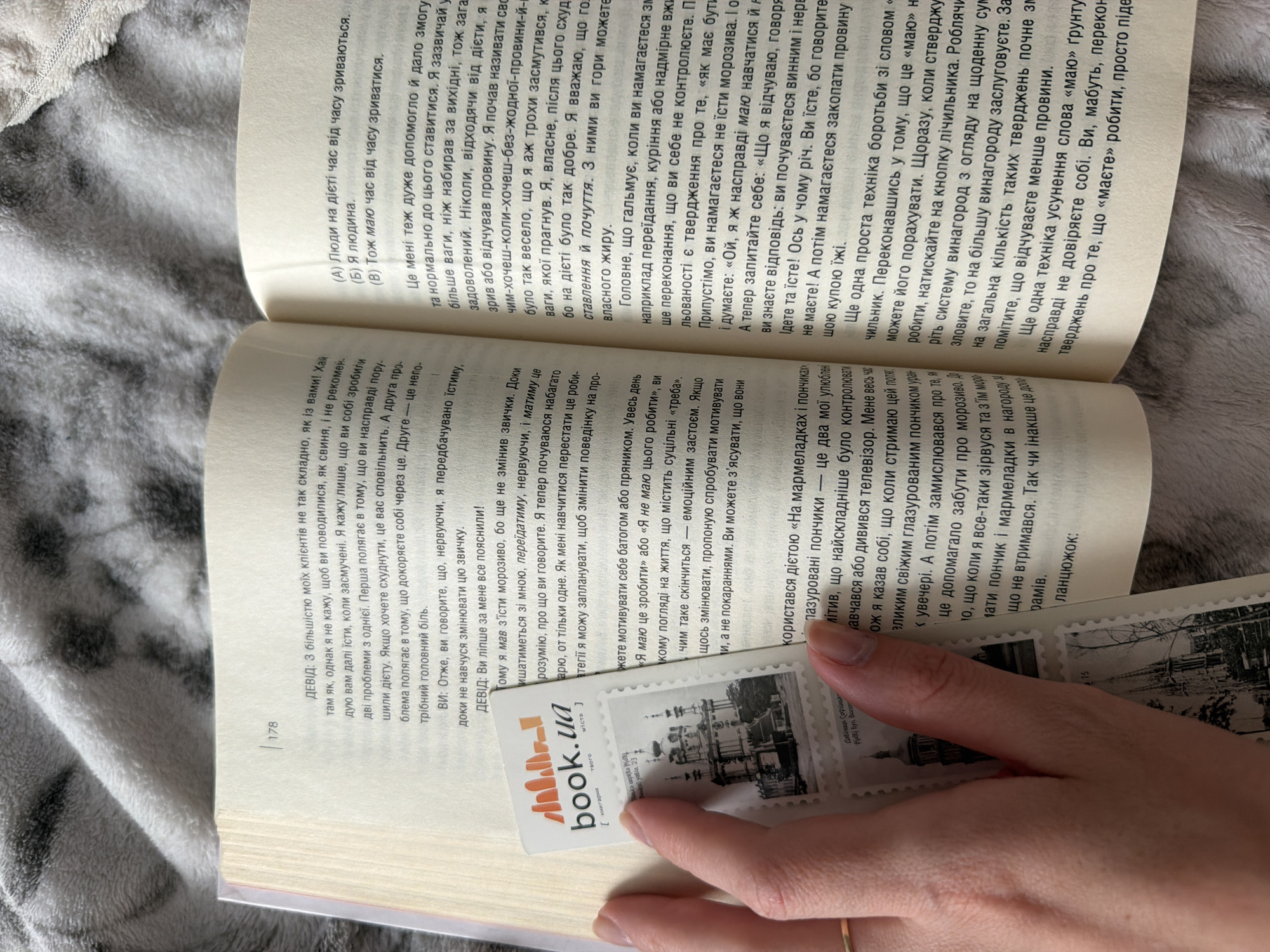 A person's hands are holding an open book on a textured grey blanket. The book is open to a page with Ukrainian text. On the left side of the book, a hand is holding a bookmark with the logo "book.ua" and an image of a church. This bookmark also features smaller images of what appear to be stamps with depictions of historical buildings. One of these stamp images shows St. Sophia Cathedral in Kyiv. The book's page number 178 is visible at the top left. The text discusses personal growth, self-discipline, and motivation.