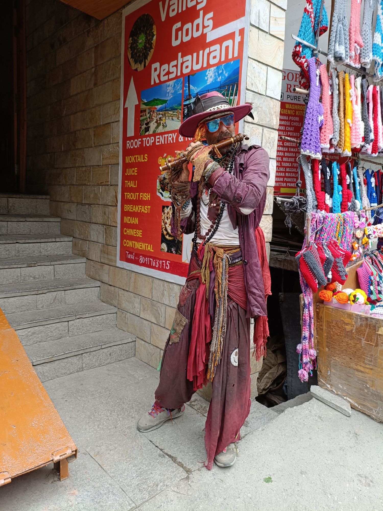 A man dressed in bohemian-style clothing, including a wide-brimmed hat, sunglasses, and layered fabrics, plays a wooden flute. He stands in front of a brick building with a red sign that reads "Valley Gods Restaurant." The sign also lists various food items and a taxi number. To the right of the man, a stall displays a variety of colorful knitted scarves and hats. The ground is paved with concrete and stone, and a set of concrete stairs is visible to the left of the man. The overall scene suggests an outdoor market or restaurant area in Vashisht, India.
