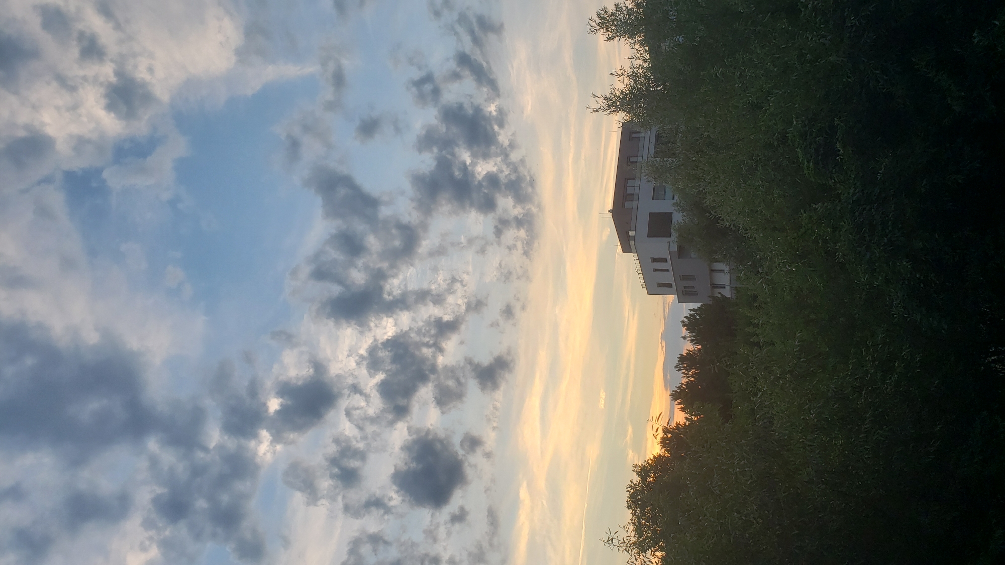 The image captures a view of a modern white building with multiple windows and balconies, situated amidst dense green foliage. The building appears to be located in a residential area, possibly on the outskirts of a city, given the presence of trees and the expansive sky. The sky is a mix of blue and grey clouds, with a warm orange and yellow hue suggesting either sunrise or sunset. This specific view is from Prešov, Slovakia.