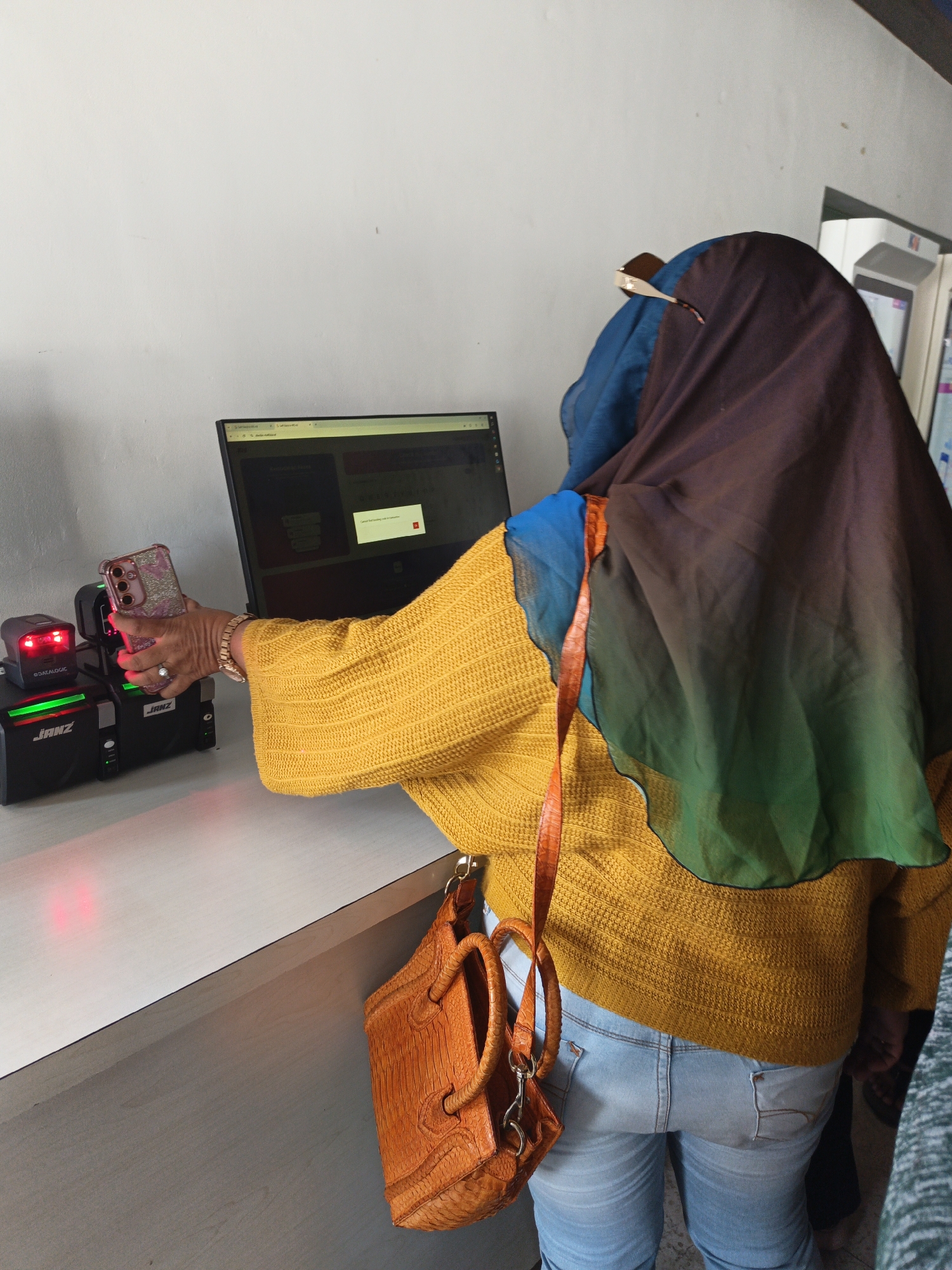 In Klaten, Indonesia, a woman wearing a yellow knitted sweater and light blue jeans, with a gradient hijab covering her head and shoulders, is shown holding up a smartphone. The phone's screen is pointed towards a scanning device. She is also wearing a small, textured orange handbag with a strap. In front of her is a computer monitor displaying a web page, and to the left of the phone, there are two black scanning devices. The scene appears to be set up on a light-colored desk against a white wall.
