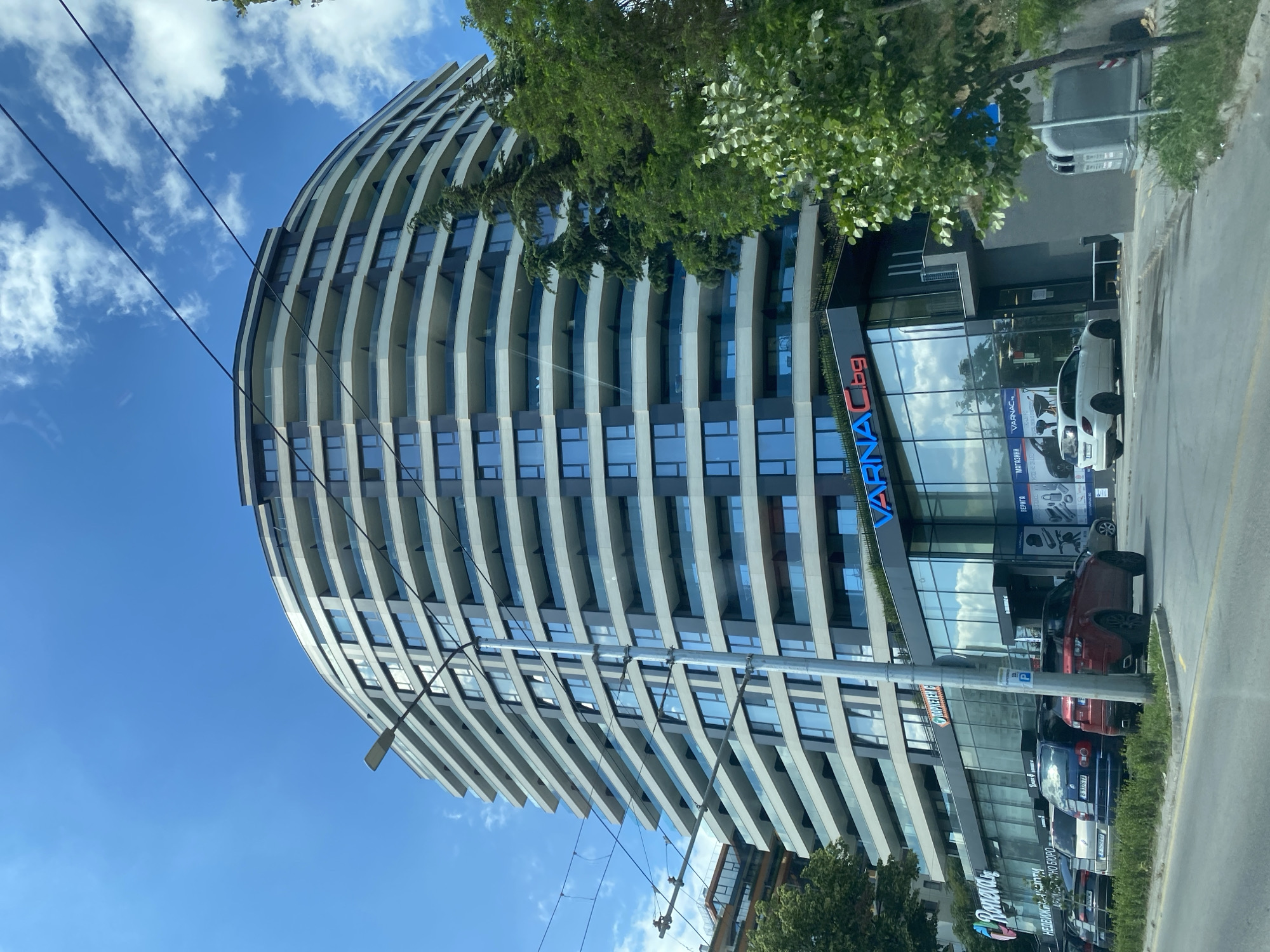 The image captures a modern, curved residential and commercial building on a bright, sunny day in Varna, Bulgaria. The building features a striking facade with alternating dark and light horizontal bands, punctuated by numerous windows and balconies. The ground floor is lined with large glass storefronts, housing businesses with signage. Visible signs include "Roneva," "VARNAC.bg," and a smaller sign with Cyrillic text indicating a store. Several cars are parked along the street in front of the building, including a red Range Rover and a white Mercedes-Benz wagon. Lush green trees and foliage surround the building, and power lines crisscross the sky, typical of an urban environment. The sky is a clear blue with scattered white clouds.