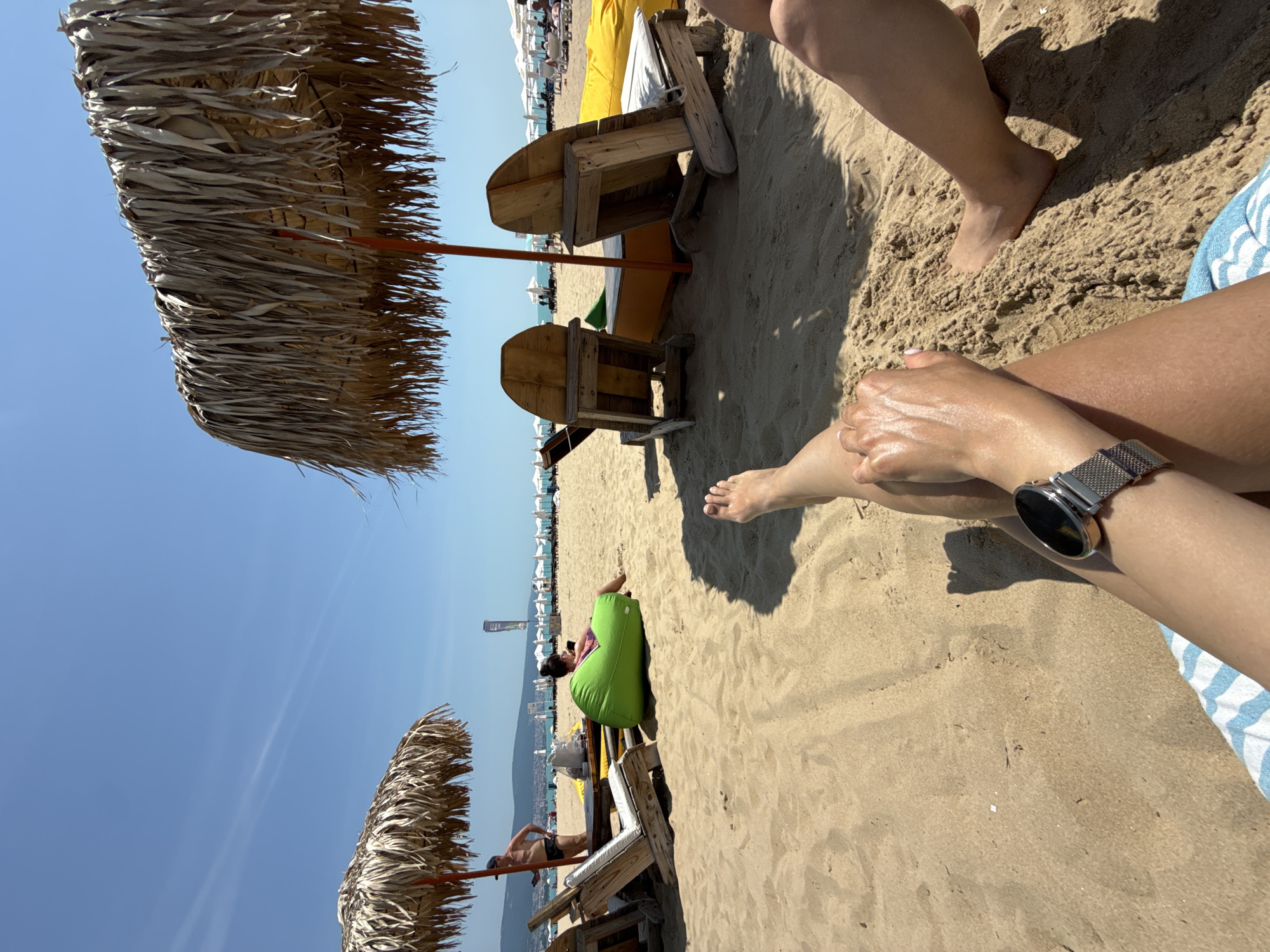 The image depicts a sunny beach scene in Nesebar, Bulgaria, viewed from a low perspective. In the foreground, the legs of two individuals are visible on light brown sand. The left leg, belonging to the photographer, wears a silver-toned smartwatch with a mesh band on the left wrist, and a blue and white striped towel is partially visible beneath. The right leg, belonging to a companion, is bare, with a foot resting on the sand.
The midground features two thatched straw parasols. The prominent central parasol has a red pole, under which two rustic wooden Adirondack-style chairs with white cushions face away from the viewer towards the sea. A small, yellow, possibly wooden, platform or table is positioned between these chairs. To the left of the central parasol, a shirtless man in black swimming shorts stands near a wooden table, looking at an object in his hand. Adjacent to him, a woman sits on a bright green bean bag chair, looking down at a device. A second thatched parasol is partially visible on the far left.
In the background, a wide sandy beach stretches into the distance, lined with multiple rows of white beach umbrellas and sun loungers. The calm, blue Black Sea extends to the horizon, where distant mountains or hills are silhouetted under a clear, light blue sky. Two faint white contrails are visible in the upper sky. Strong shadows cast by the parasols and objects indicate direct sunlight, likely midday to early afternoon.