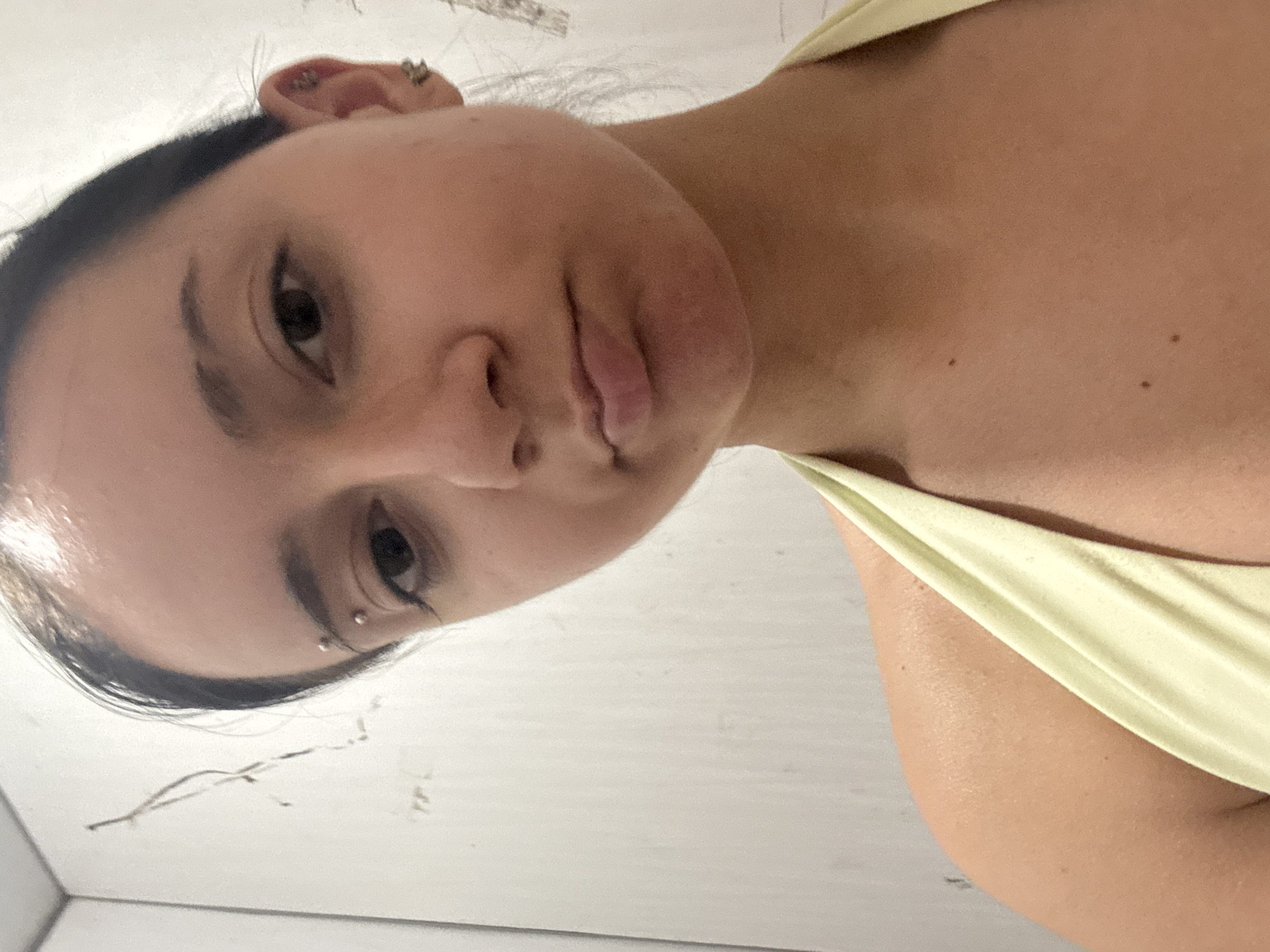 An upward-facing, medium shot shows a young woman from the chest up against a white, textured wall. She has dark hair pulled back, dark eyes, and a light complexion. A small silver hoop is in her eyebrow, and she is wearing a pale yellow halter top. She has a neutral expression and her lips are slightly pursed. Her left ear, visible on the right side of the frame, has two small silver earrings. The background wall is plain and lightly textured with faint vertical lines and what appear to be scuff marks or shadows.