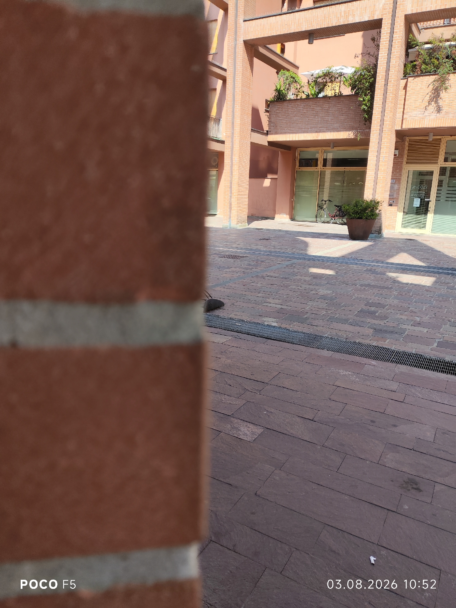 In a sunlit outdoor courtyard paved with reddish-brown rectangular stones, a brick building with an arched facade and balconies adorned with greenery is visible. A bicycle is parked near a store entrance. A dark grate runs along the ground. The foreground is dominated by a blurred brick pillar. The lower left corner displays "POCO F5" and the lower right corner shows the date "03.08.2026" and time "10:52".