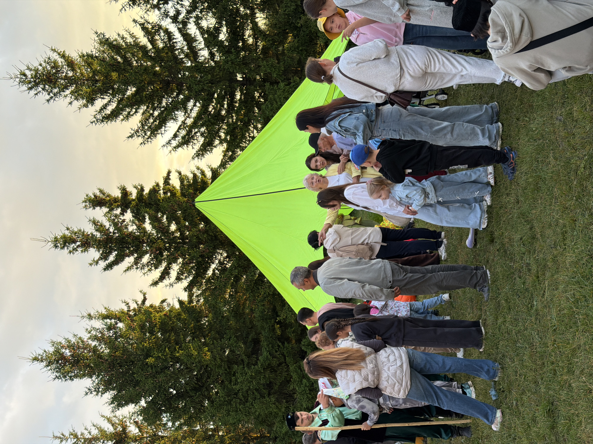 A diverse group of people are gathered in an outdoor setting, with a vibrant lime green tent as the focal point. The tent is large and triangular, positioned in the center of the frame. A man in a green tunic and dark hat, holding a wooden stick, is interacting with a woman. Several other individuals, including women, men, and children, are standing around, engaged in conversation or observing. The background features tall, dark green coniferous trees, suggesting a forest or park environment. The sky above is a light yellow, indicating either sunrise or sunset. The ground is covered in green grass, with some dry leaves scattered about. The overall atmosphere appears to be a casual outdoor gathering or event.