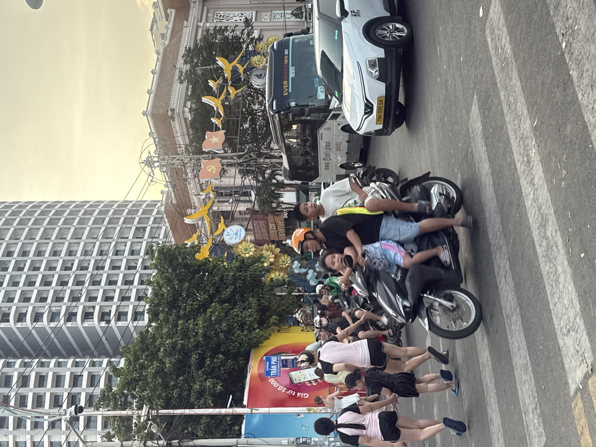 In Nha Trang Ward, Vietnam, a street scene unfolds with pedestrians and vehicles at a crosswalk. A family of four, two adults and two children, are on a scooter. The adult male at the front wears an orange helmet and a black shirt, while the adult female behind him wears a pink and black top and black shorts. A young boy in a blue and pink shirt and shorts sits between them, and a younger child in a blue and pink patterned outfit is positioned in front of the boy.
To the left, three females are walking away from the camera. One, in a black and pink top and black shorts, holds hands with a young girl in a black dress. Another female, wearing a black and pink top and black shorts, walks ahead. A fourth female, in a white top and black shorts, is further ahead.
In the background, a white bus with "EVER GREEN NEW 2025" visible on its front is present. A white SUV with license plate "79H-095.68" is also stopped at the crosswalk. Tall buildings, adorned with decorative yellow bird and sunflower motifs, line the street. Electrical wires crisscross above. The sky is a pale yellow, suggesting either dawn or dusk.